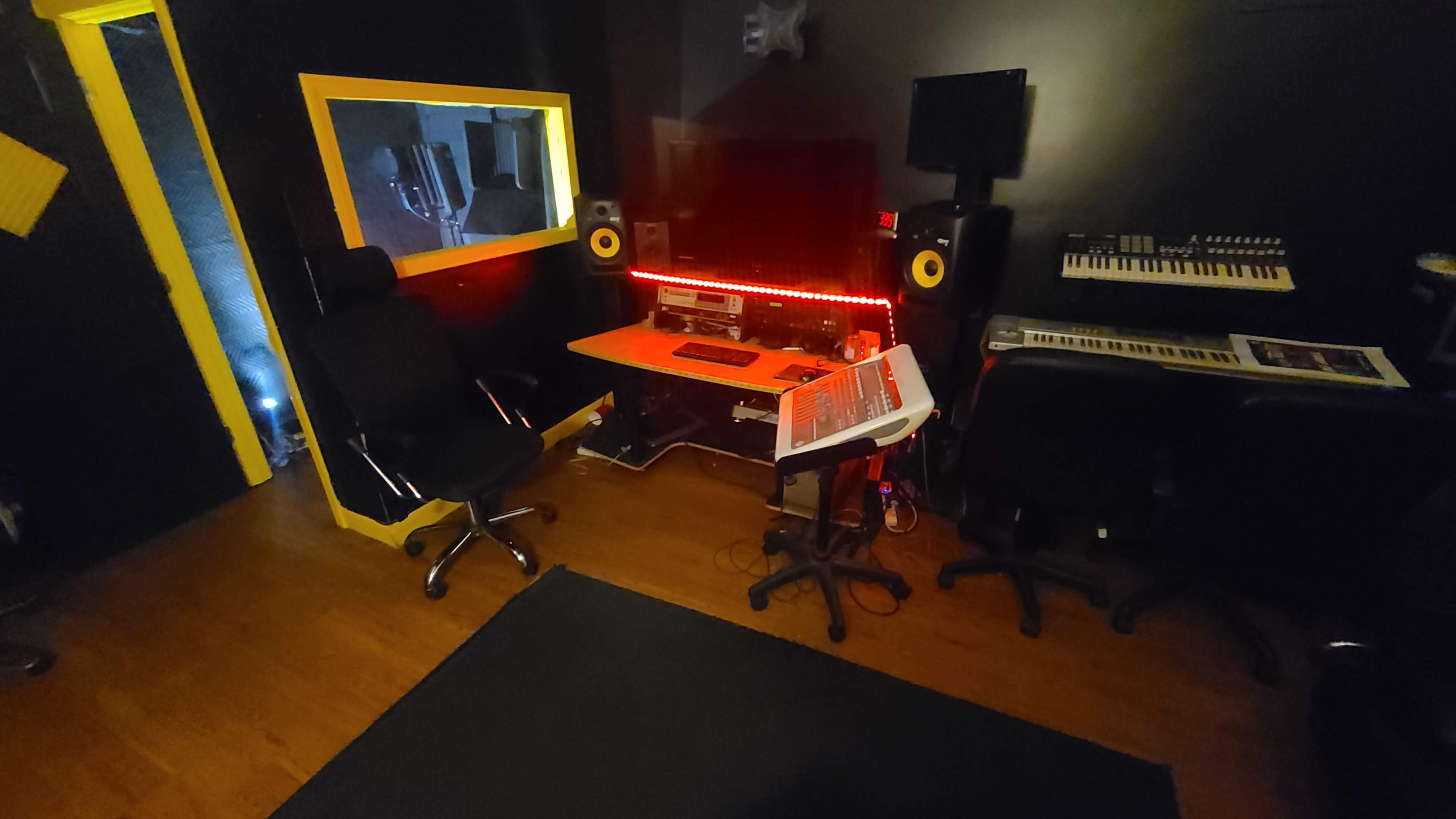 A dimly lit music studio featuring a large monitor, a keyboard, two studio monitors, and two office chairs, with red LED lighting along the desk.