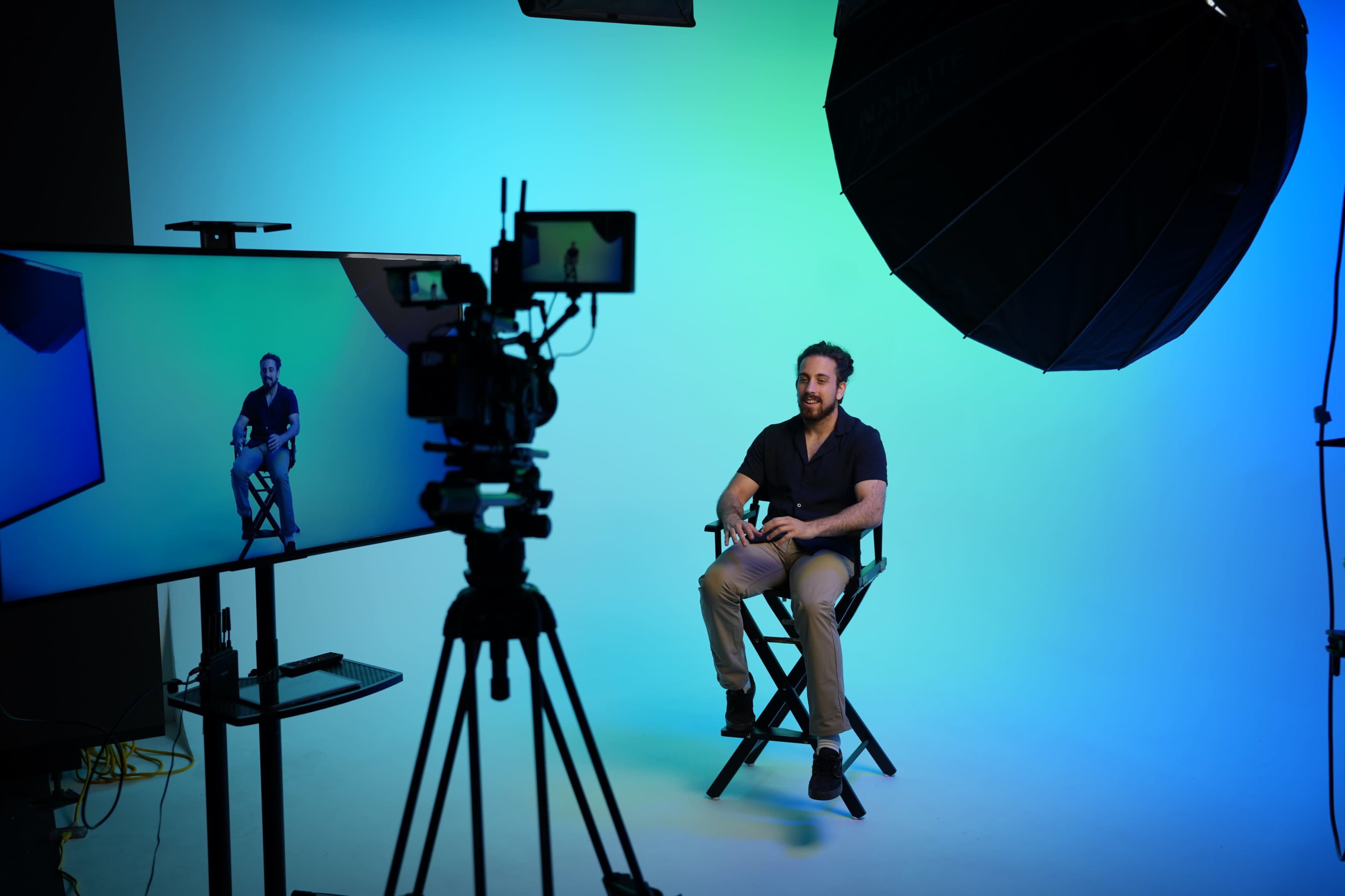 A man sits on a director's chair in front of a camera setup with a large softbox light and a monitor displaying his image in a studio with a gradient background.