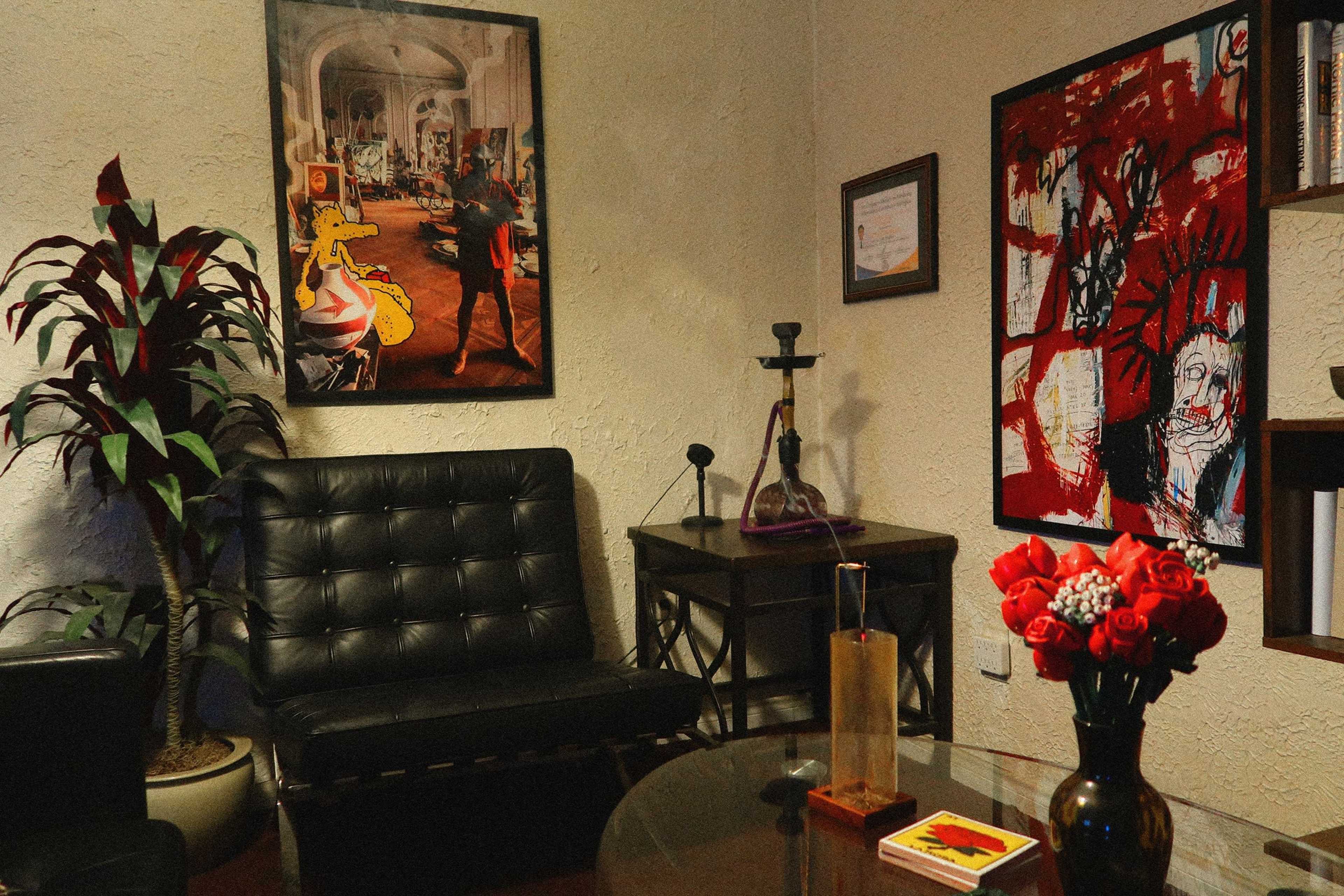 The image shows a cozy room featuring a black sofa, artwork on the walls, a coffee table with a vase of red roses, and a decorative plant in the corner.