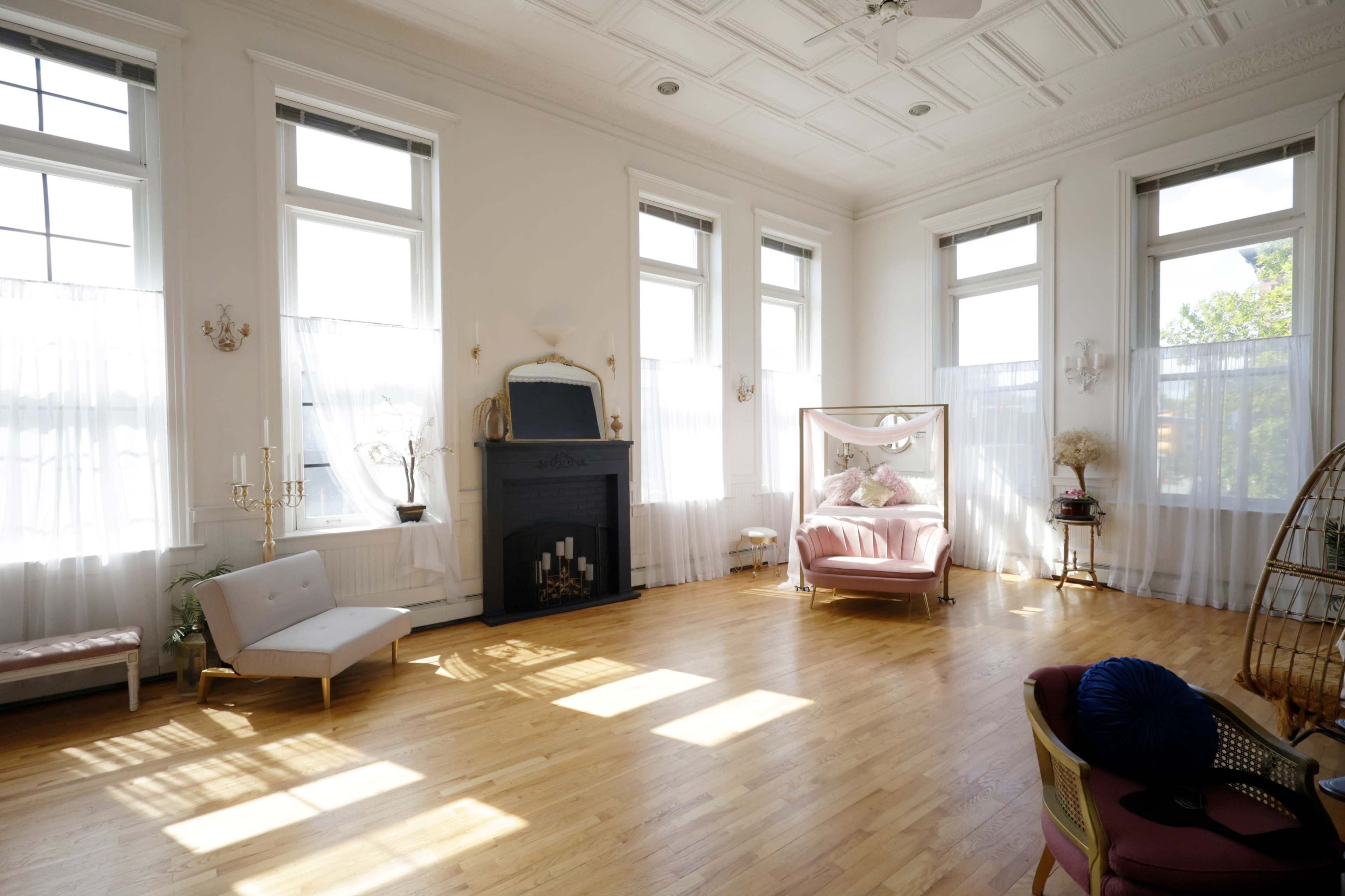 The image features a spacious, bright room with multiple windows, a fireplace, and a pink armchair, complemented by light wooden flooring.