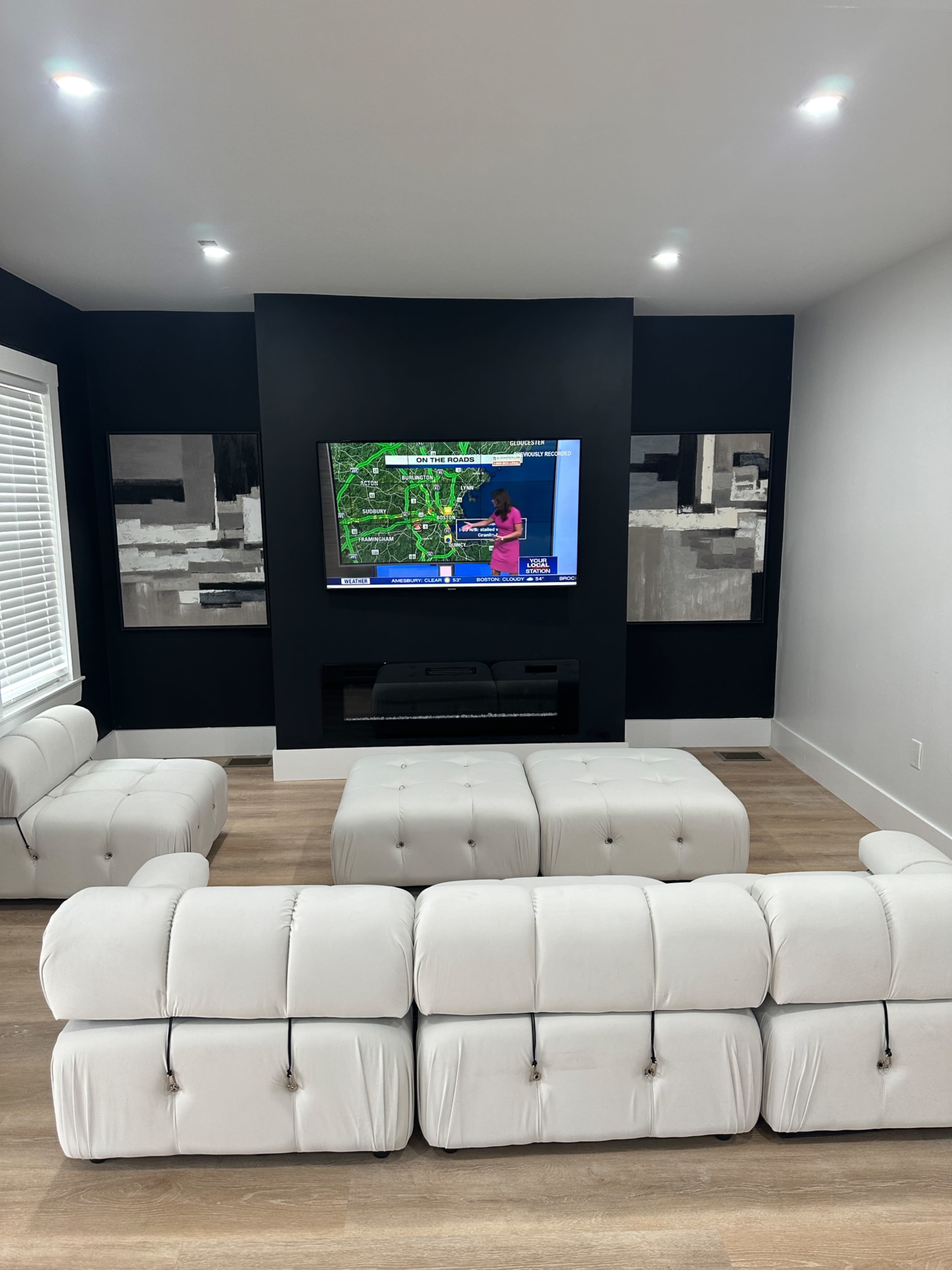 A modern living room with a white sectional sofa facing a mounted TV on a black wall, flanked by two abstract art pieces.