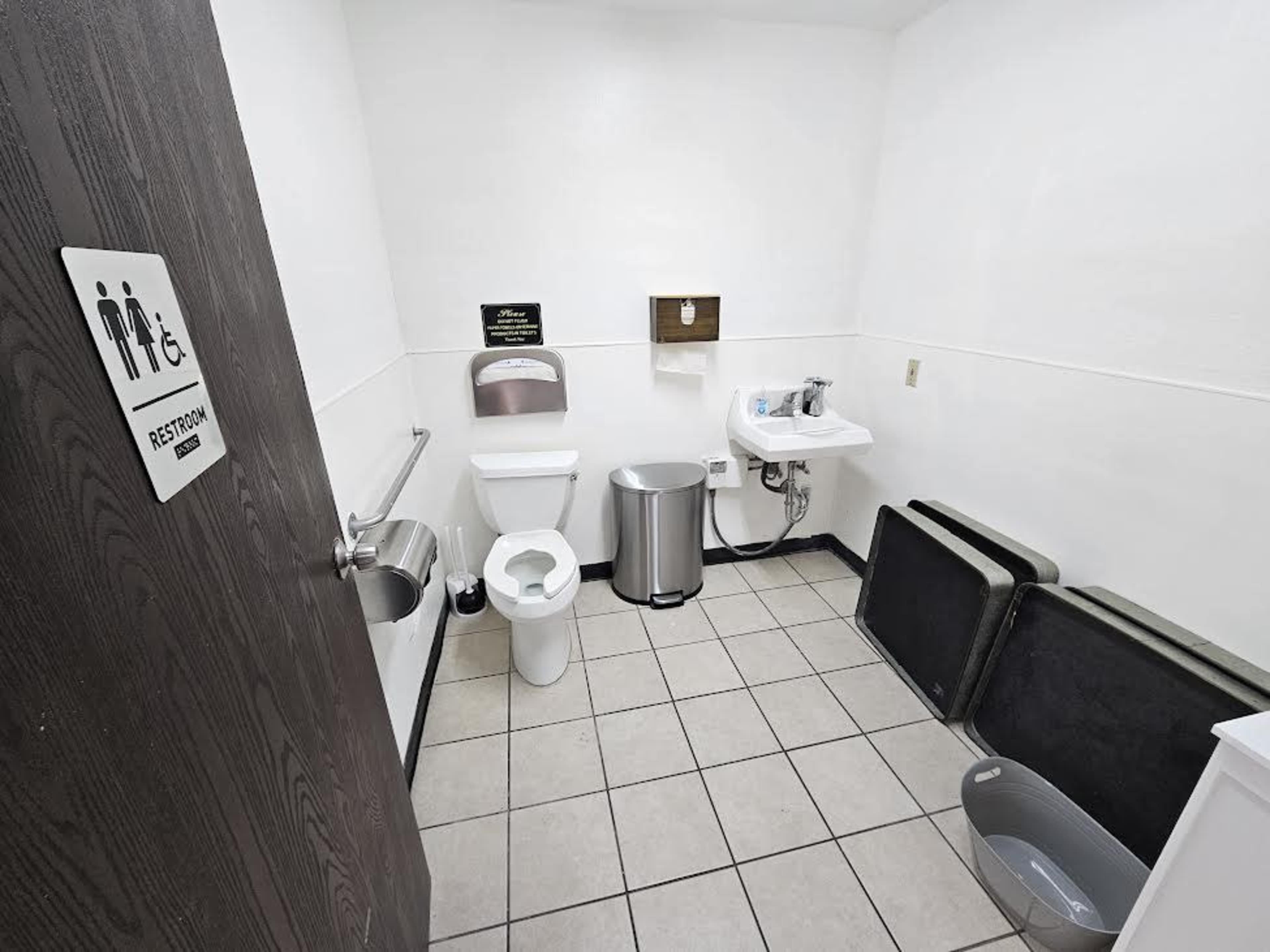 The image shows a small, clean restroom with a toilet, sink, hand dryer, trash can, and a couple of folding tables against the wall.