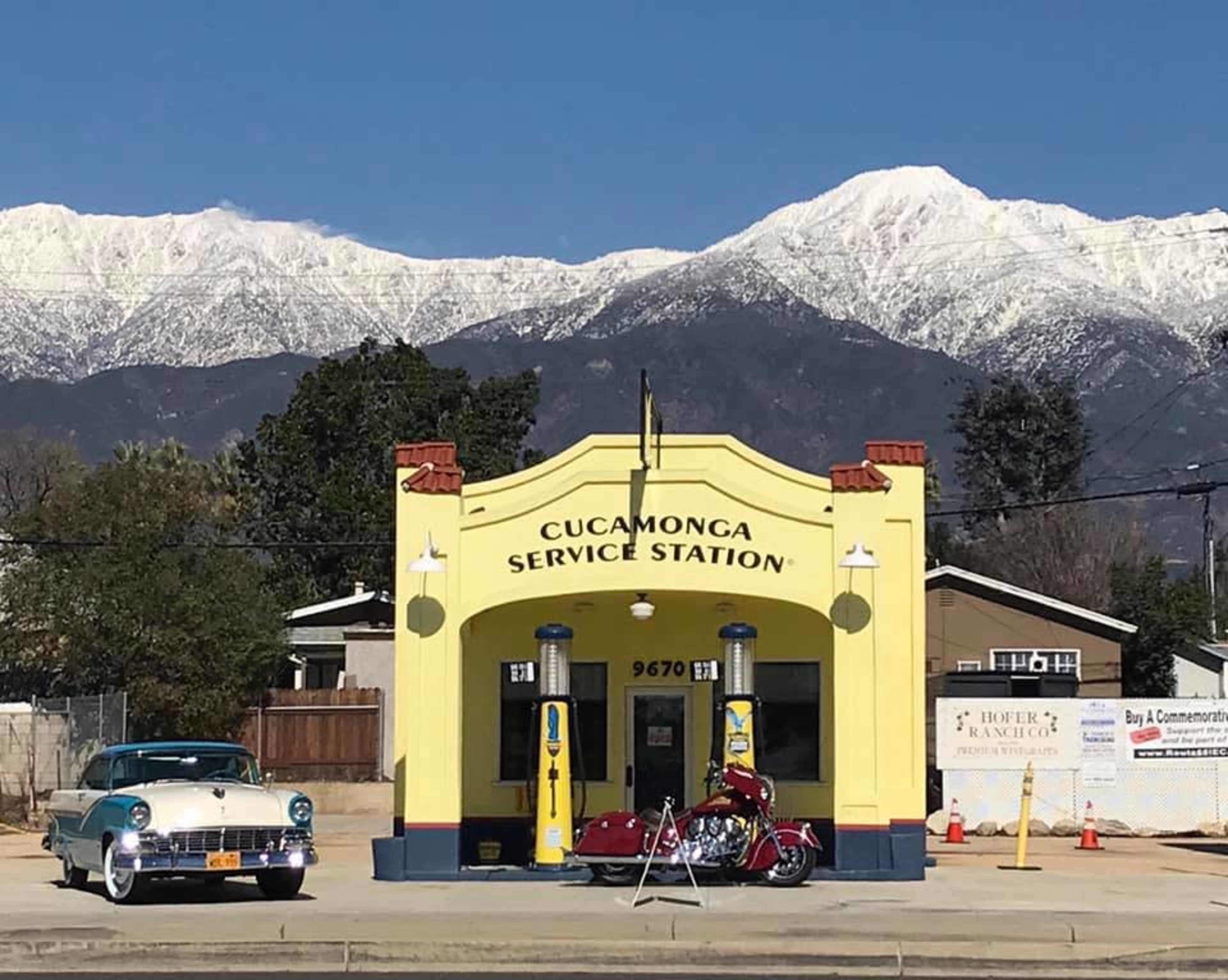 A vintage car is parked in front of a bright yellow service station with snowy mountains in the background.