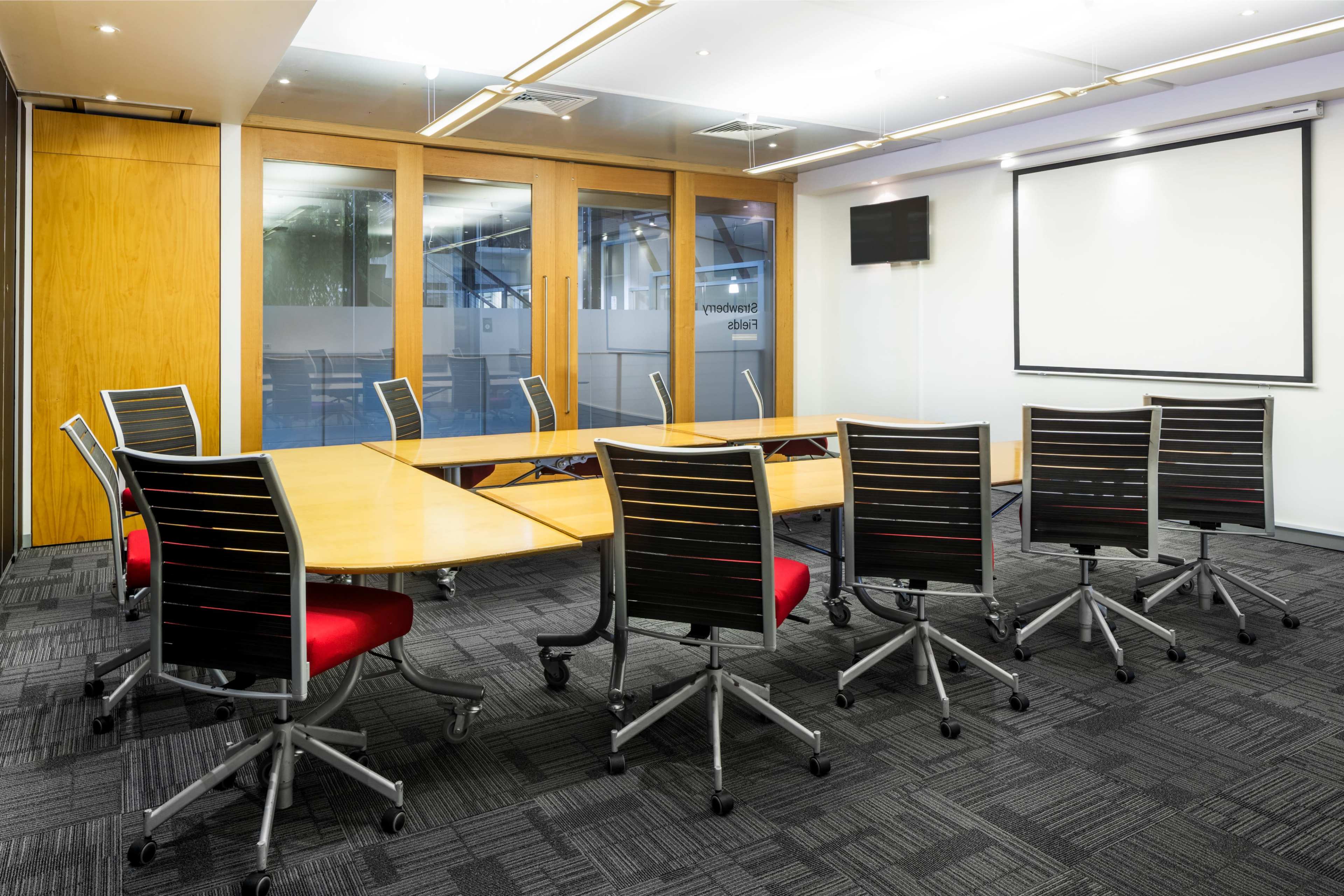 A modern conference room features a large, curved wooden table surrounded by black and red rolling chairs, with glass walls and a projection screen at one end.