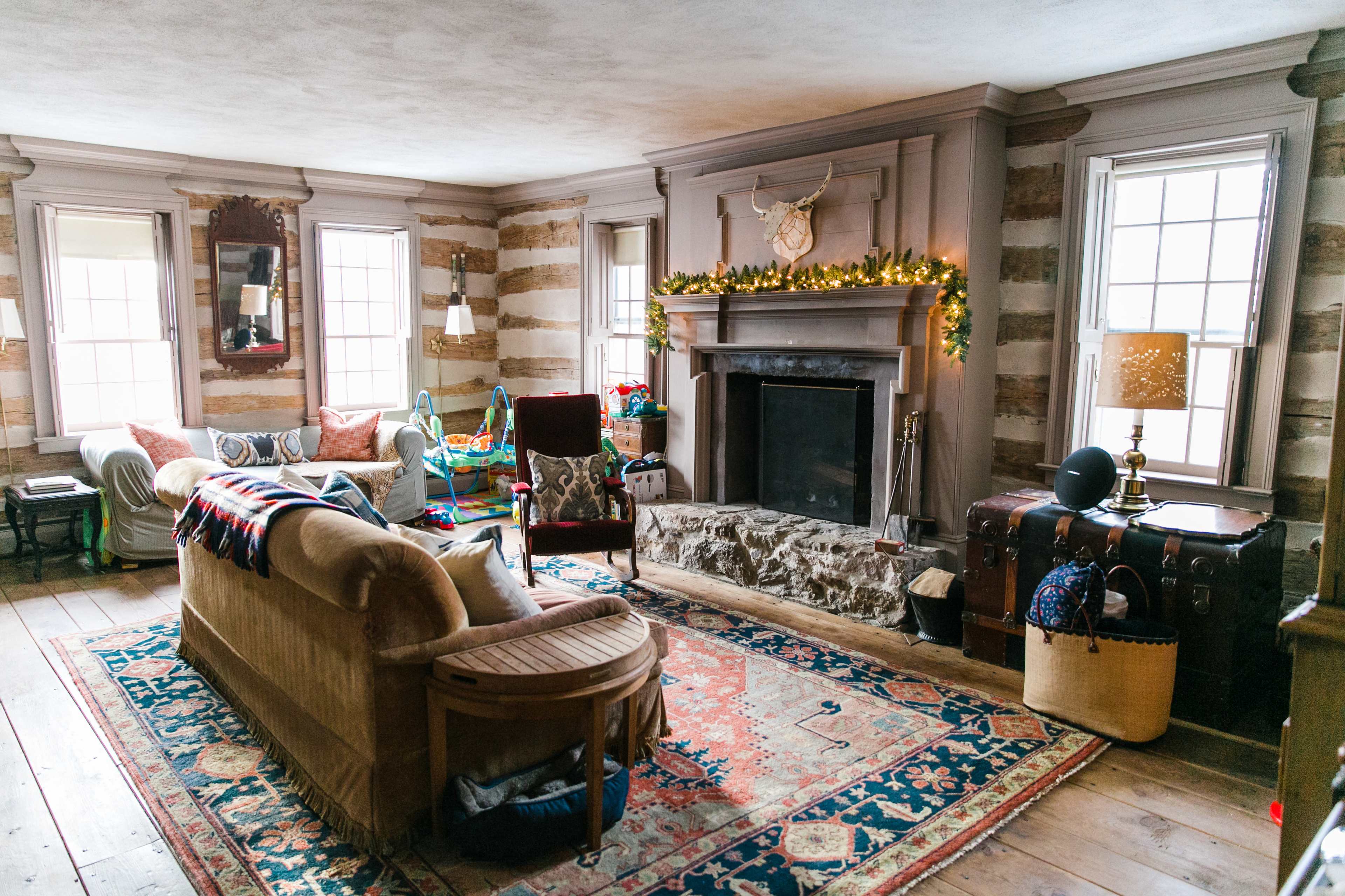 The living room features a stone fireplace, wooden floors, and a mix of vintage and modern furnishings, with large windows letting in natural light.