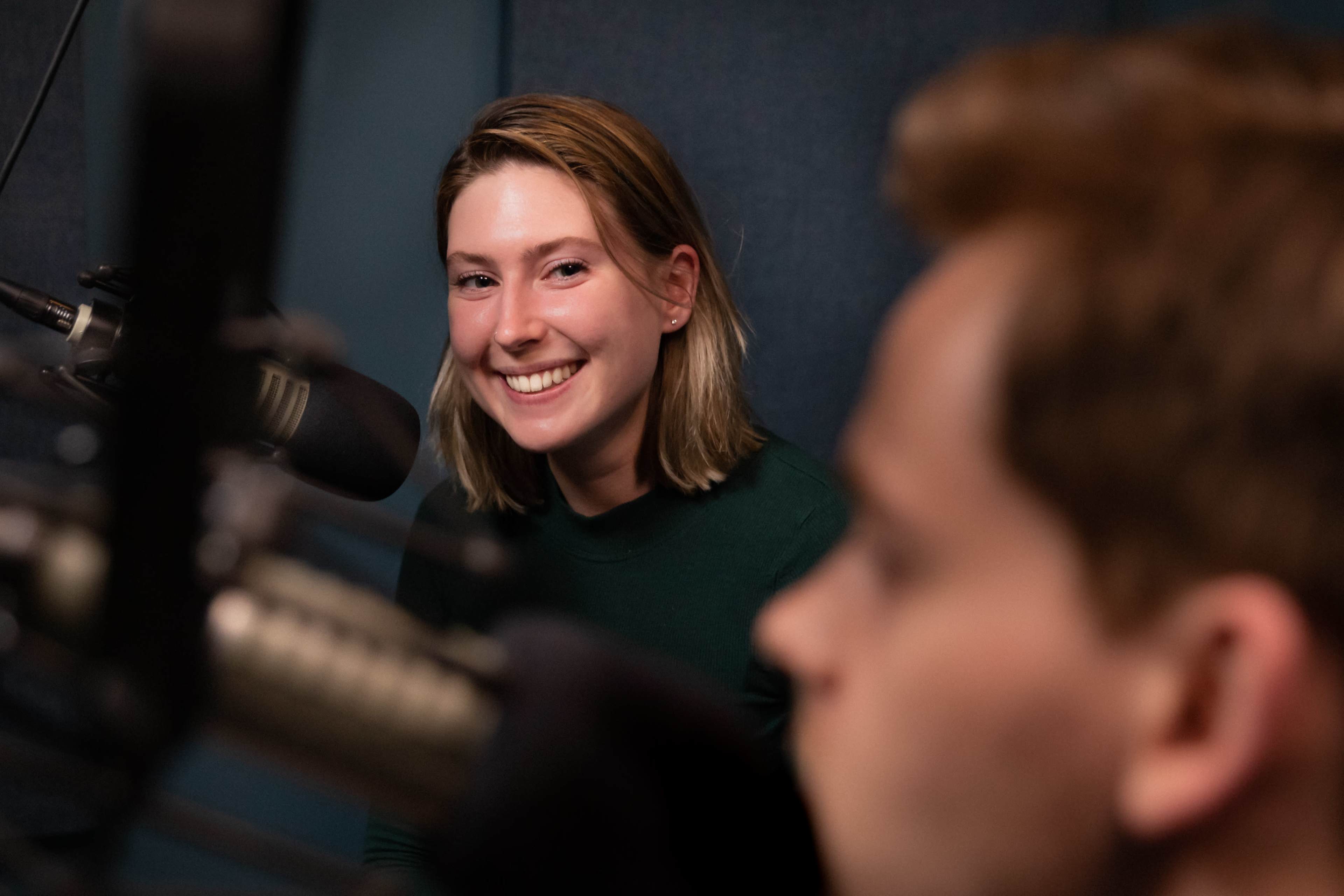 A woman smiles while sitting in front of a microphone, with a man slightly blurred in the foreground.
