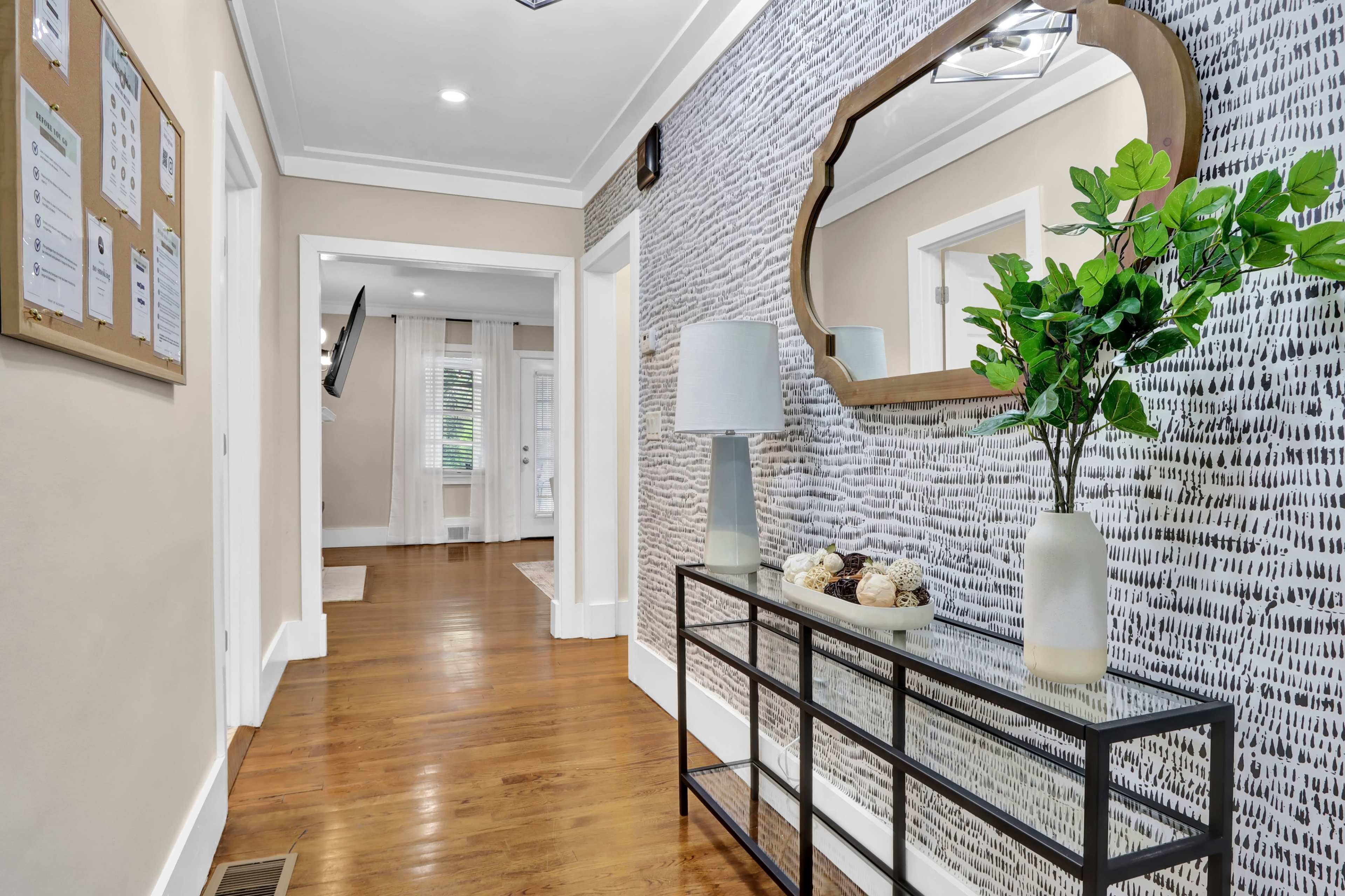 A hallway with wooden flooring, a console table topped with decorative items, a mirror on the wall, and a patterned wall treatment.