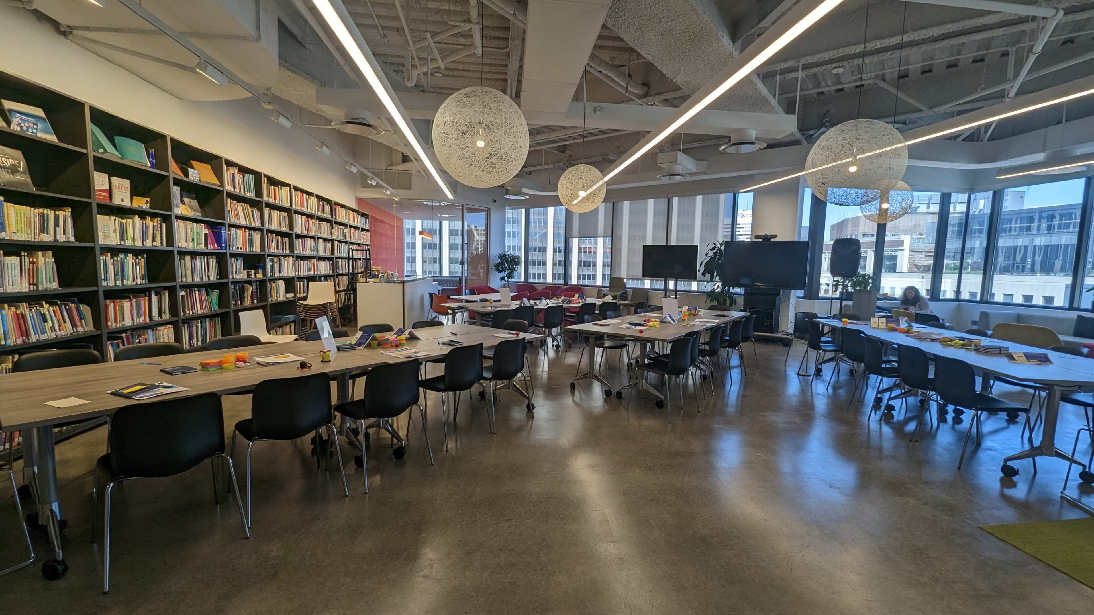 A modern meeting space with long tables arranged in a U-shape, surrounded by shelves filled with books and large windows letting in natural light.