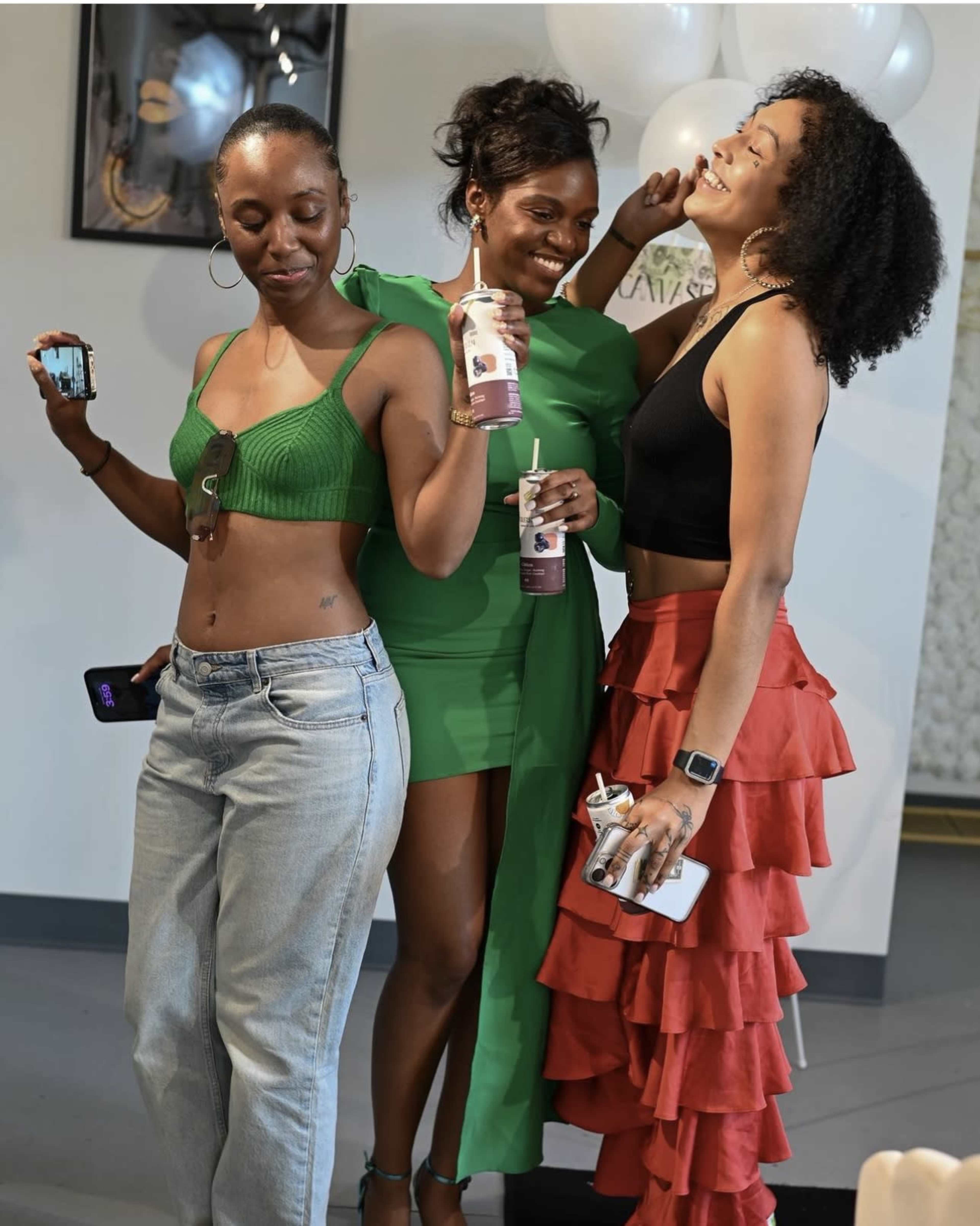 Three women pose together in a brightly lit room, each holding drinks and smiling.