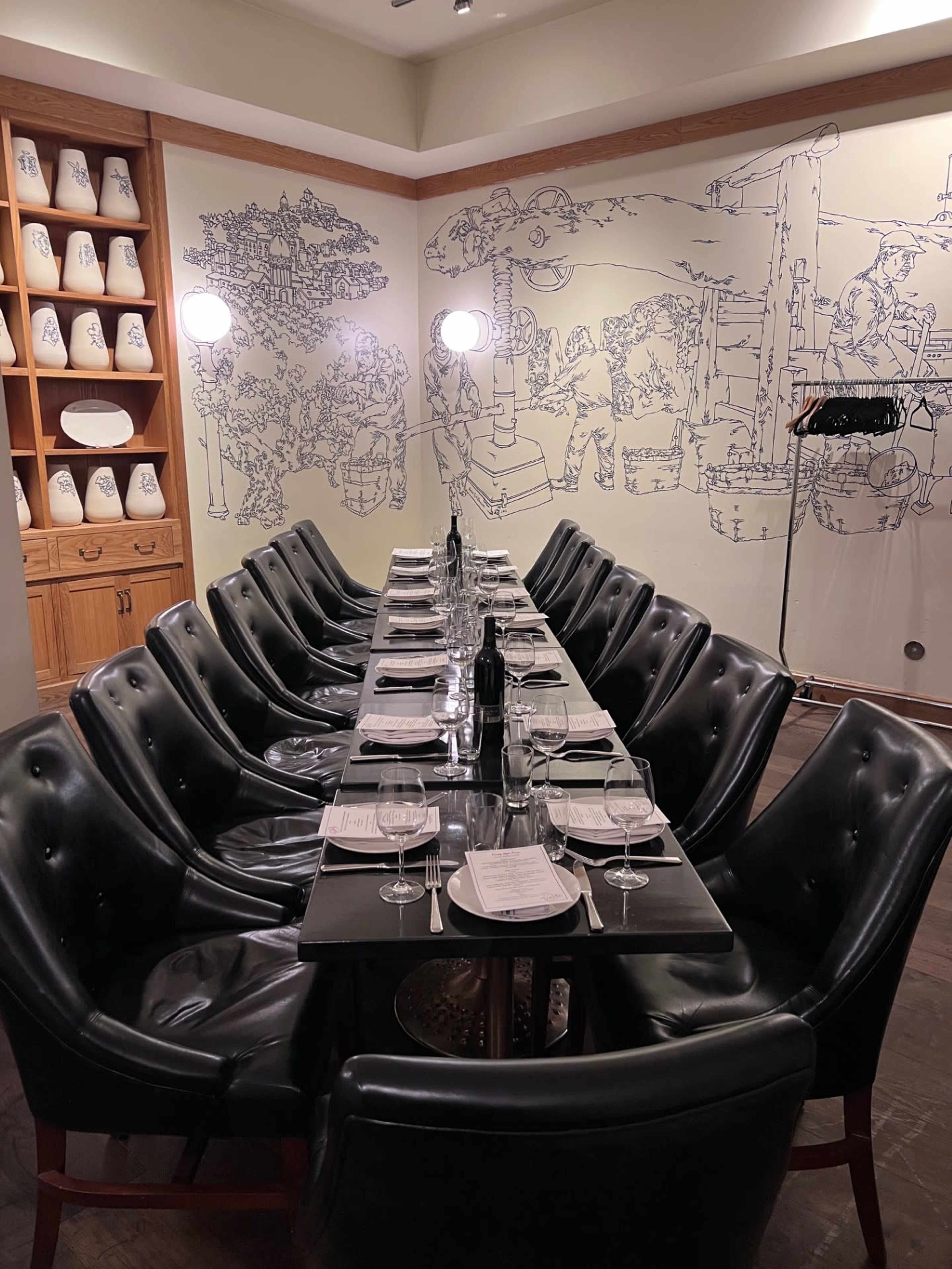 A long table is set for dining in a room featuring a decorative mural and shelves with ceramic items.