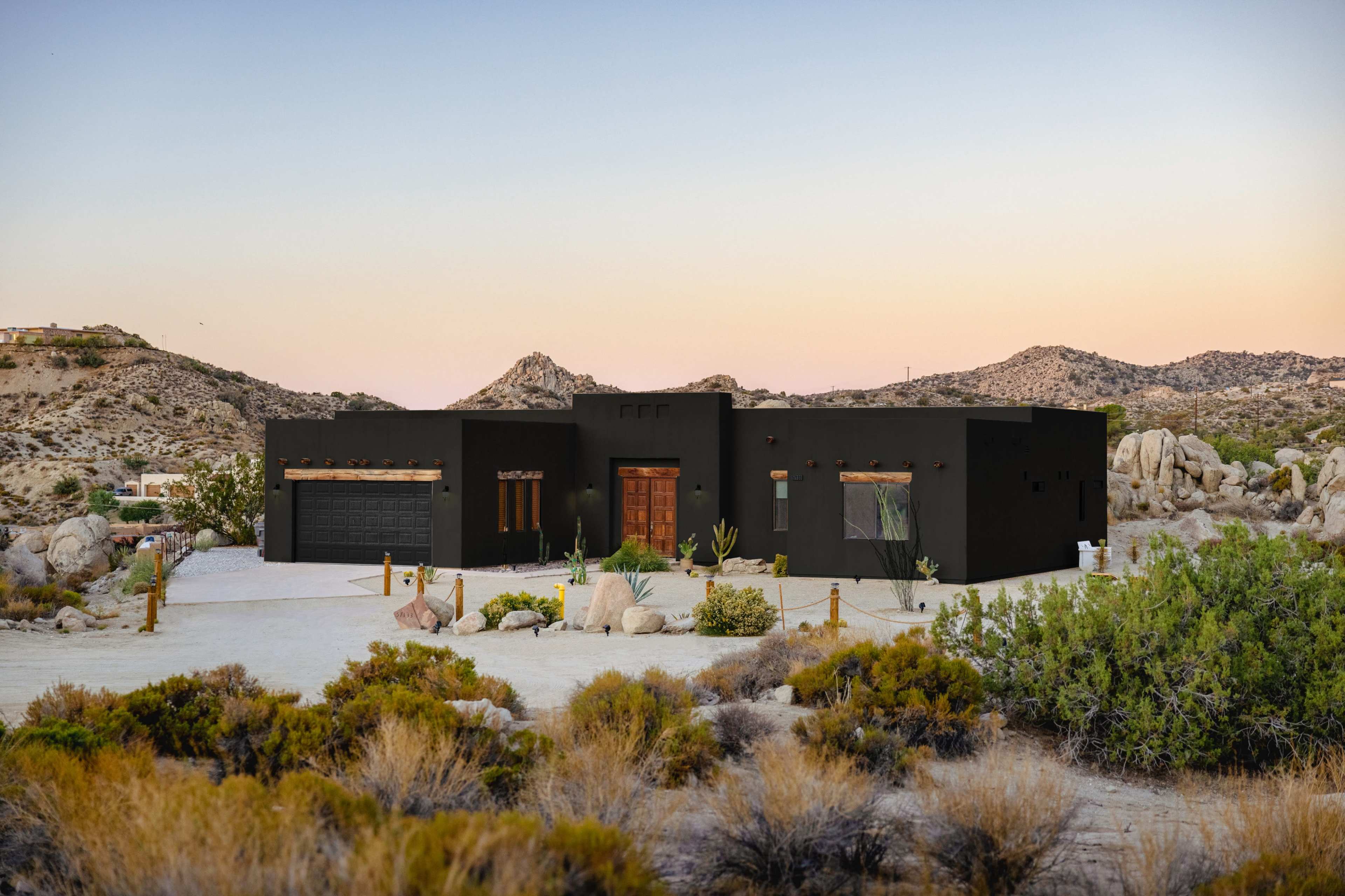A modern black house is situated in a desert landscape surrounded by rocky hills and sparse vegetation.