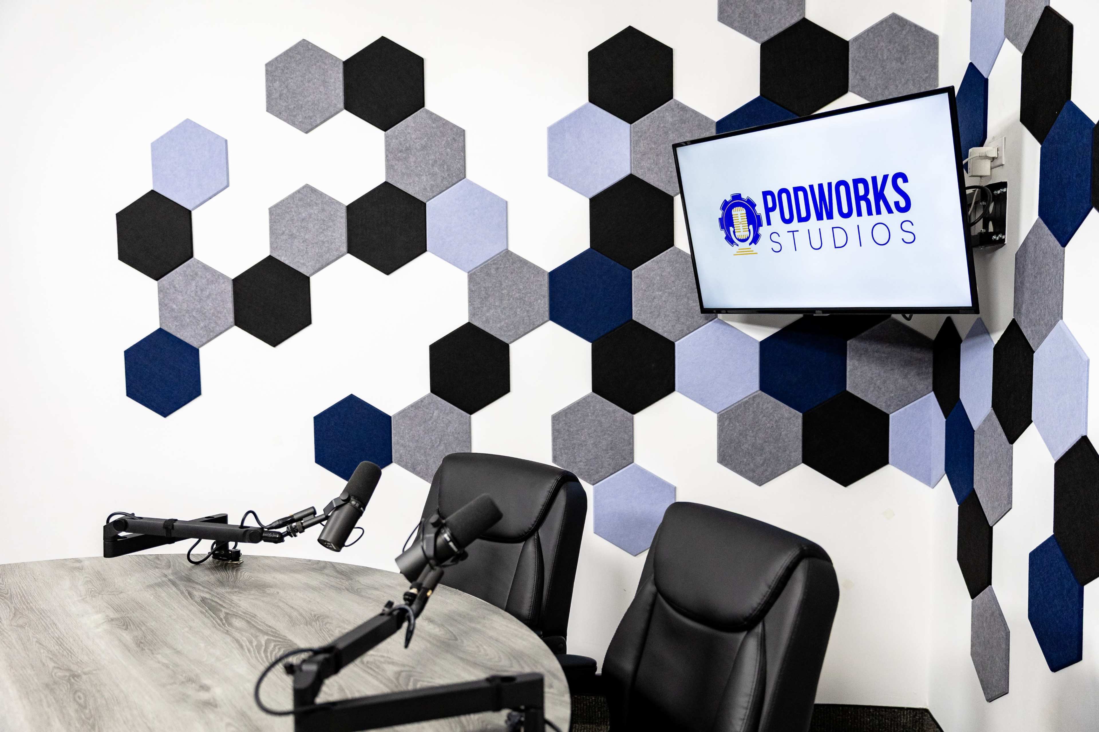 The image shows a podcast recording studio featuring a round table, two black chairs, and a wall decorated with hexagonal sound panels in various shades of gray and blue, along with a monitor displaying the "Podworks Studios" logo.