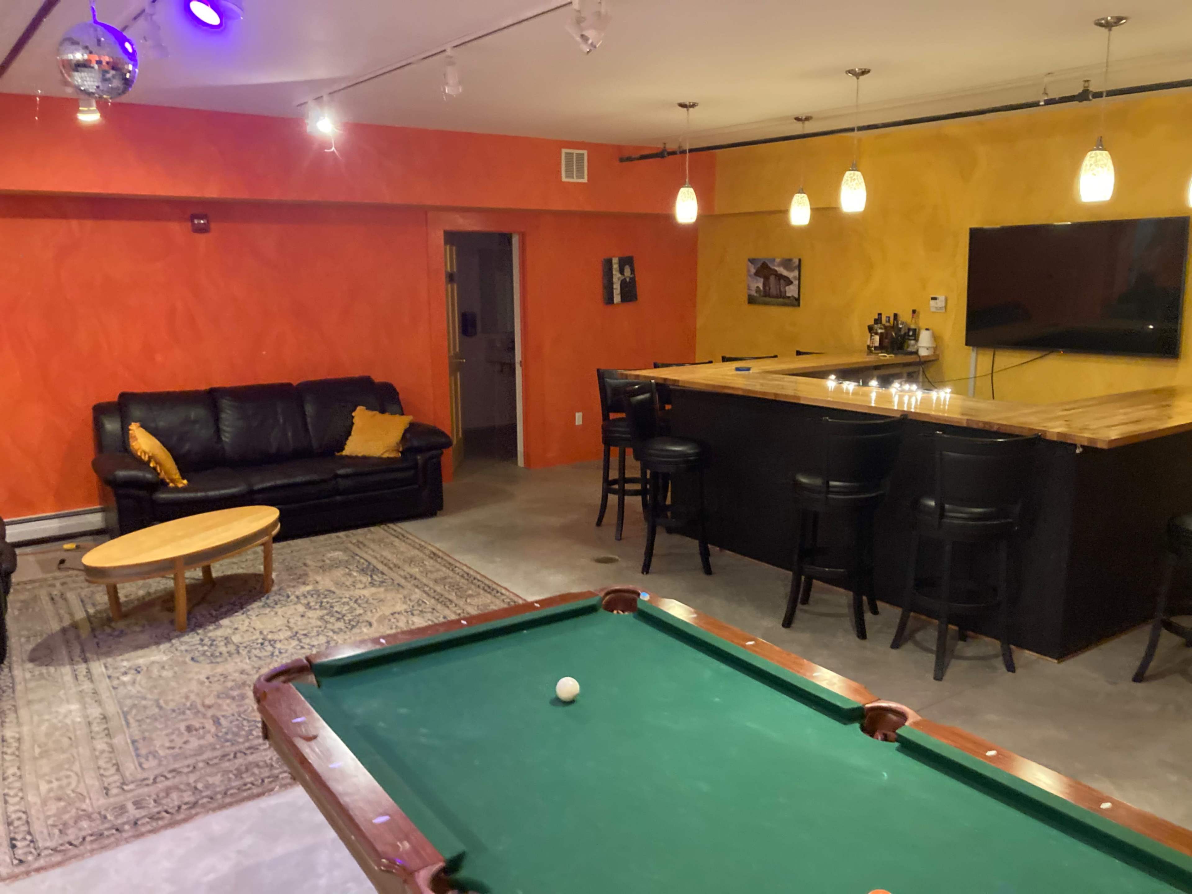 A recreational room featuring a pool table, a bar area with stools, a leather sofa, and brightly colored walls.