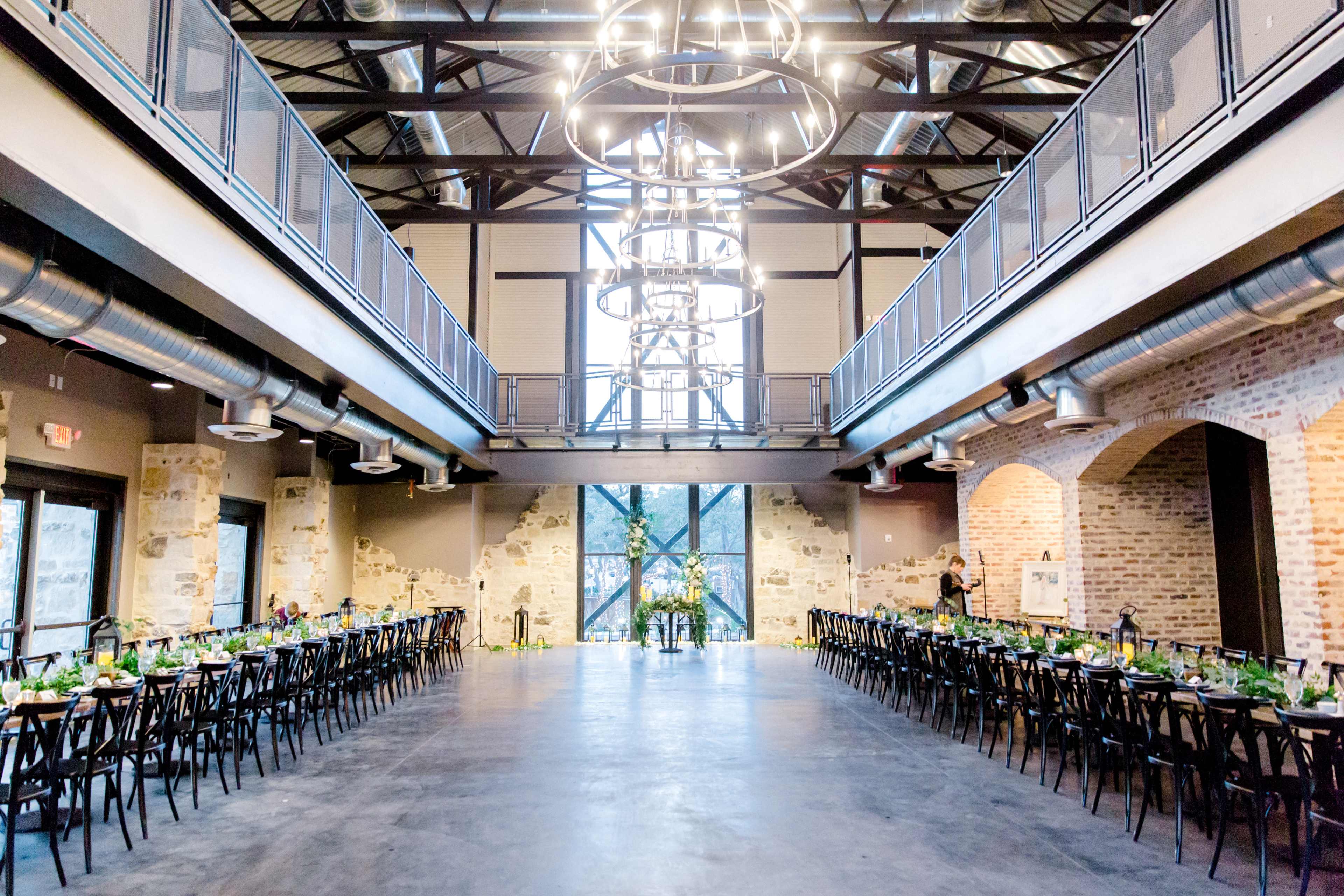 The image shows a spacious event venue with a long dining table set in the center, surrounded by large windows and industrial-style architecture.