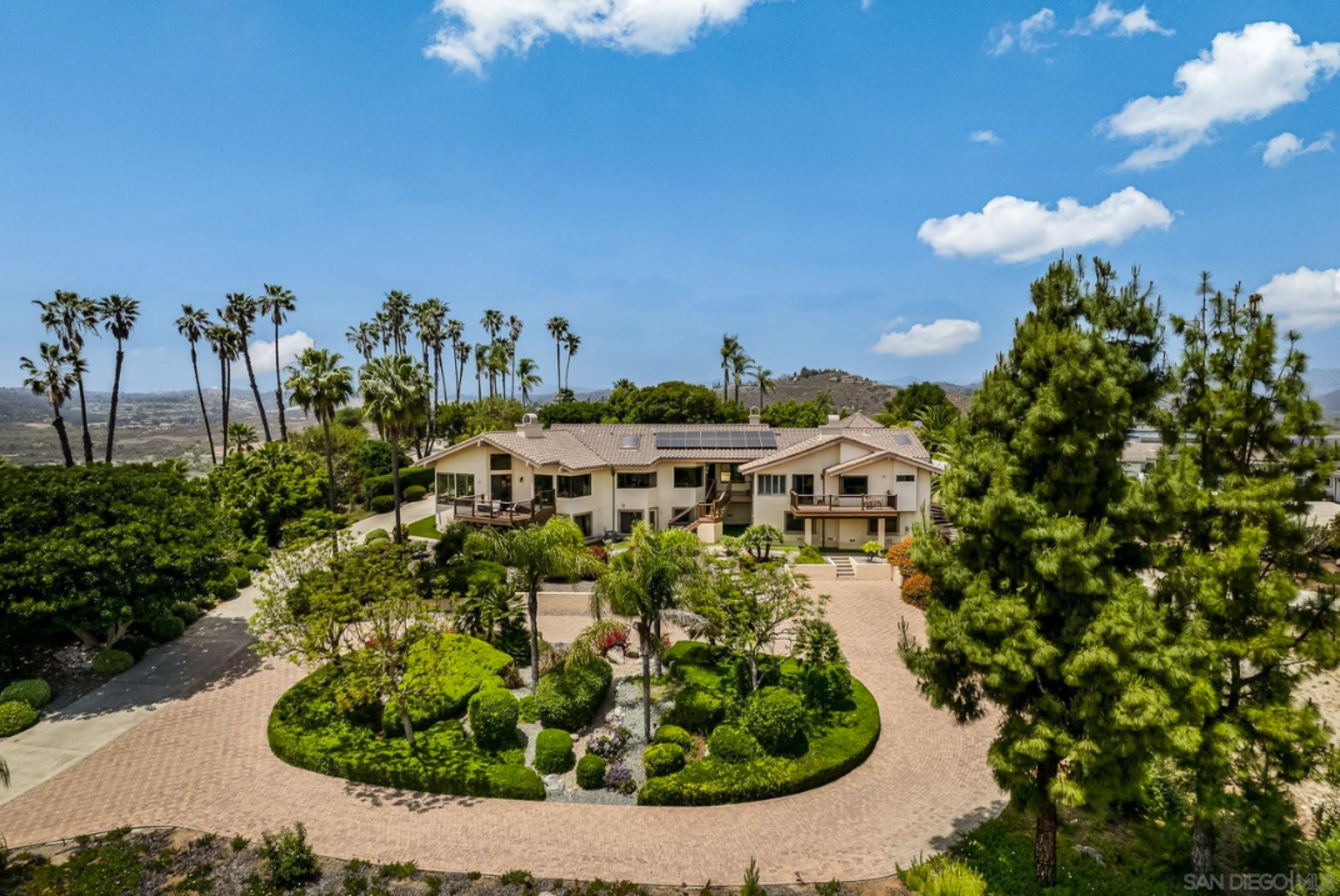 The image shows a large two-story house surrounded by landscaped gardens and palm trees, with a paved driveway leading up to the entrance.