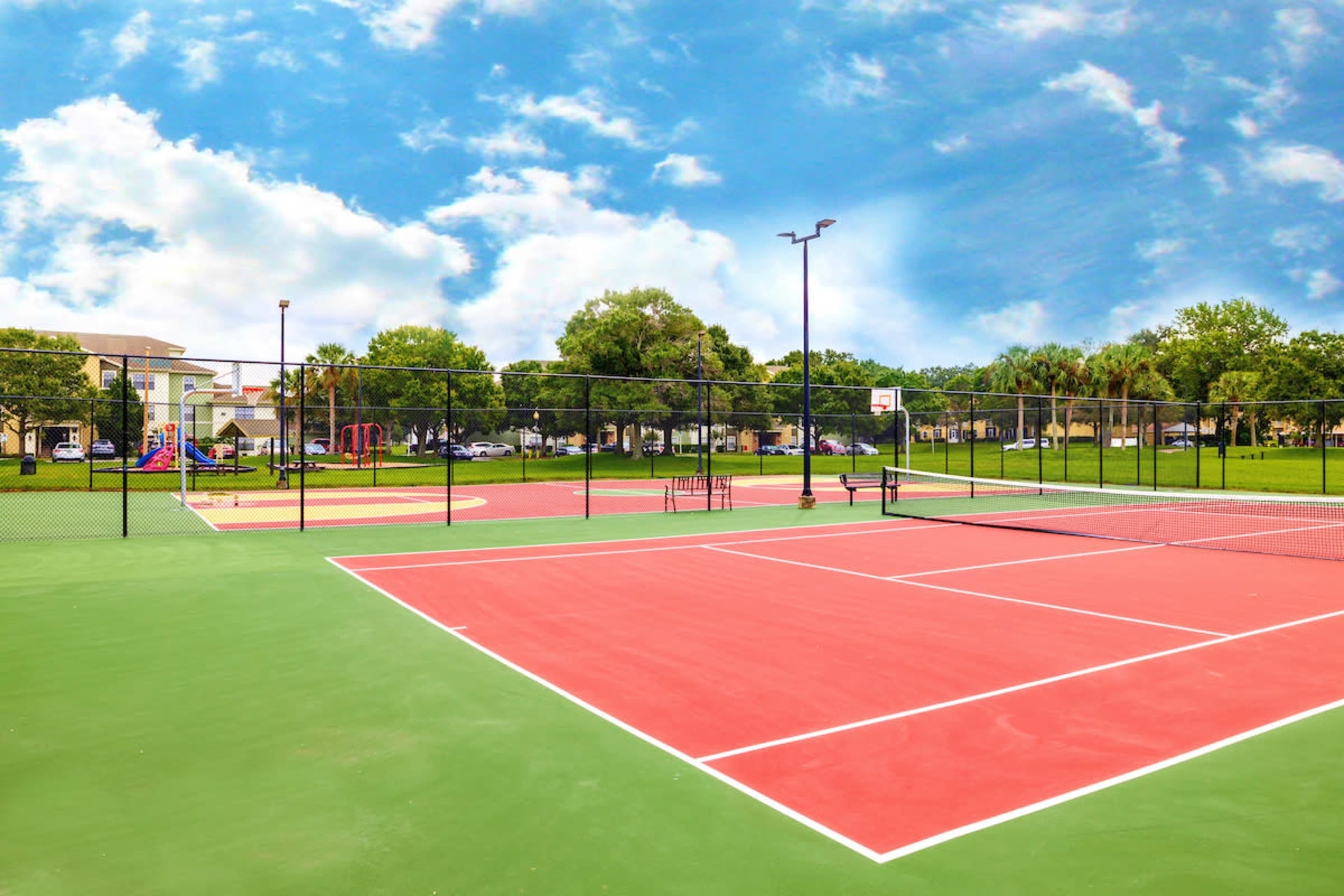 A pair of tennis courts surrounded by trees and a playground in a residential area, under a blue sky.