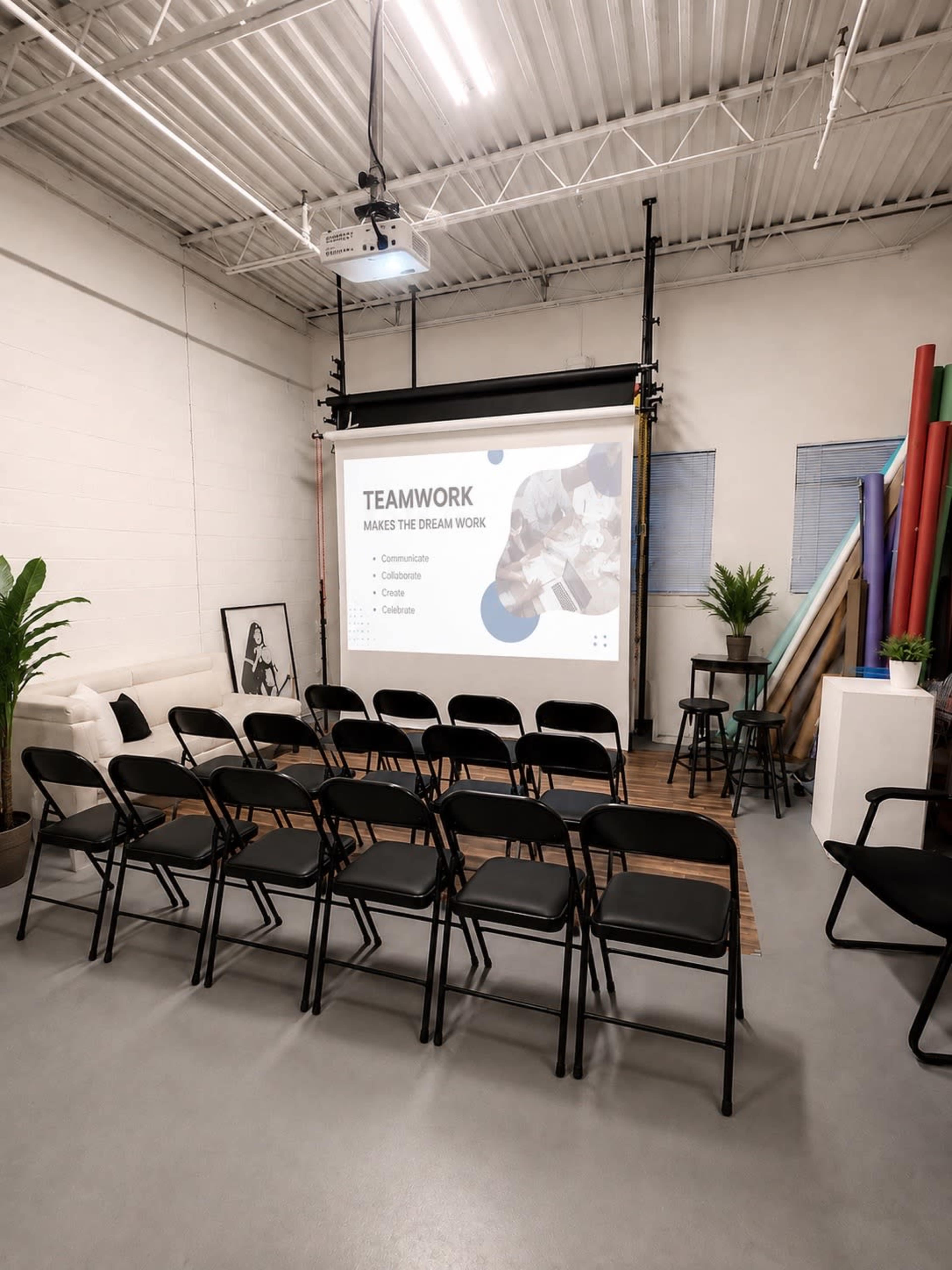 The image shows a meeting room with rows of black folding chairs facing a projection screen displaying the words "TEAMWORK MAKES THE DREAM WORK," along with a seating area featuring a couch and potted plants.