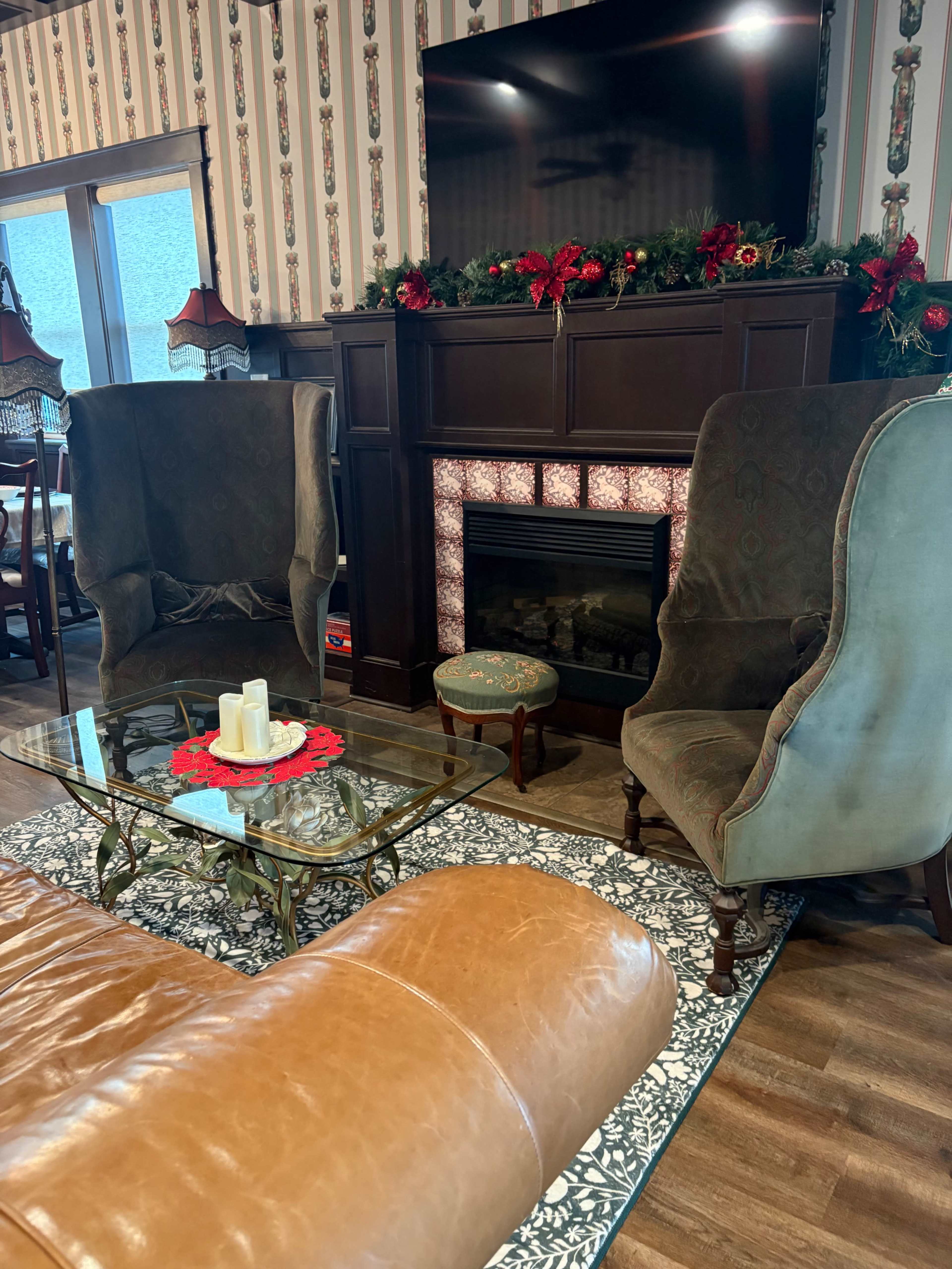 The room features a fireplace with a TV above, two wingback chairs, a glass coffee table with candles, and decorative elements like a floral rug and holiday greenery.