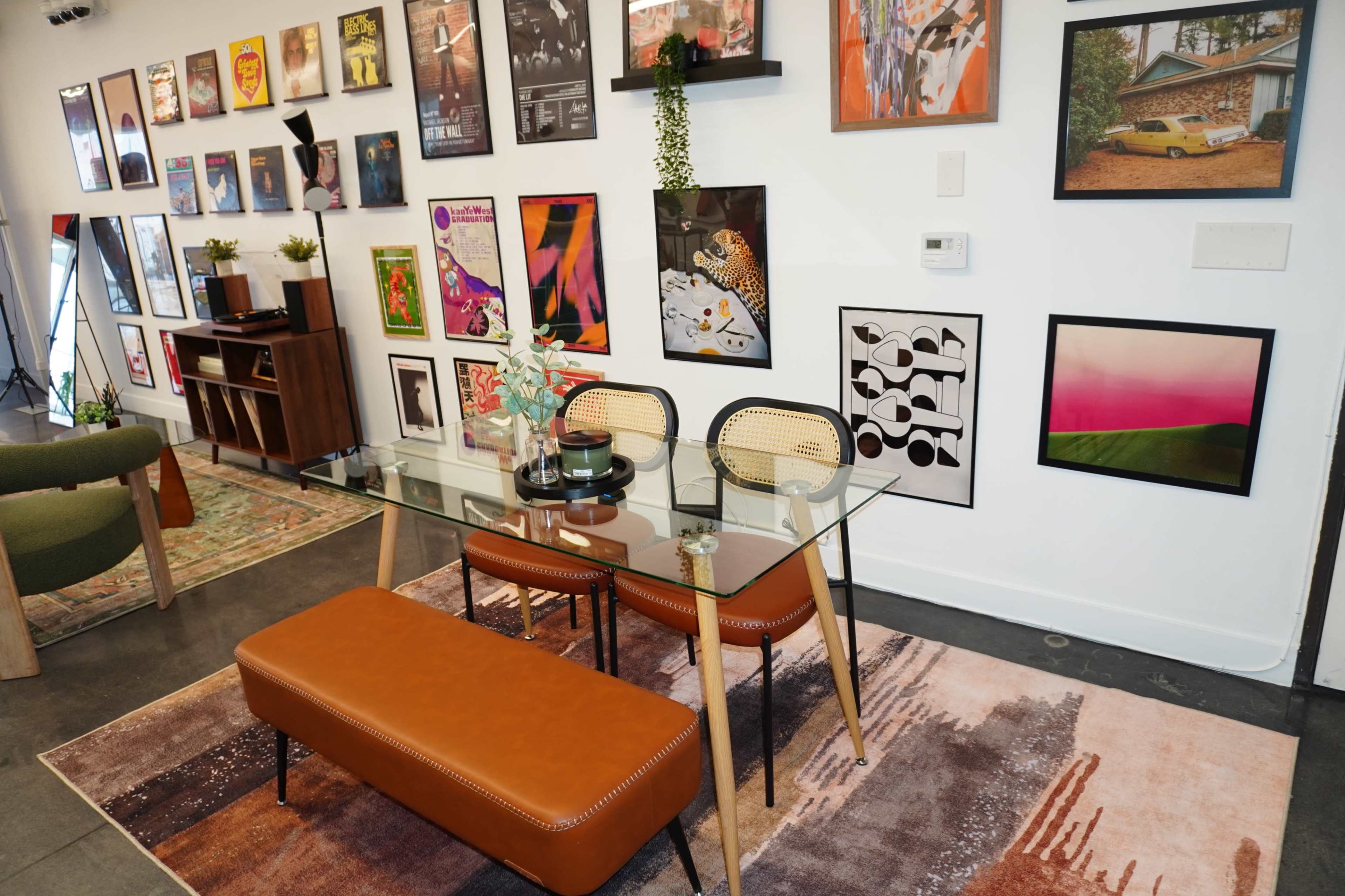 The image shows a modern dining area with a glass table, two tan leather benches, and a wall adorned with various framed posters and artworks.
