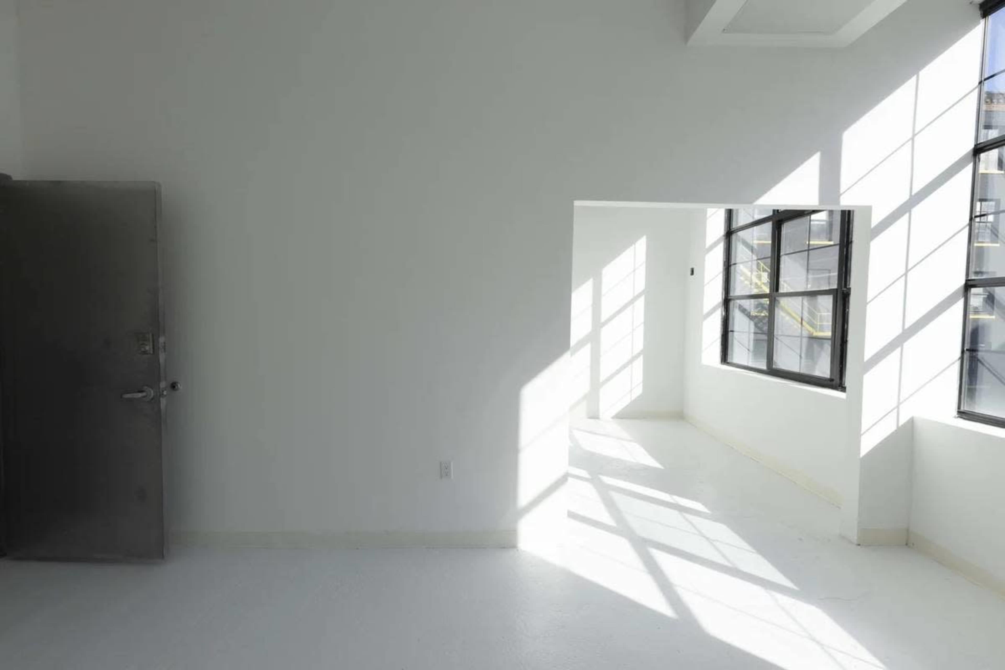 A spacious, empty room with white walls and large windows allowing sunlight to cast shadows on the floor.