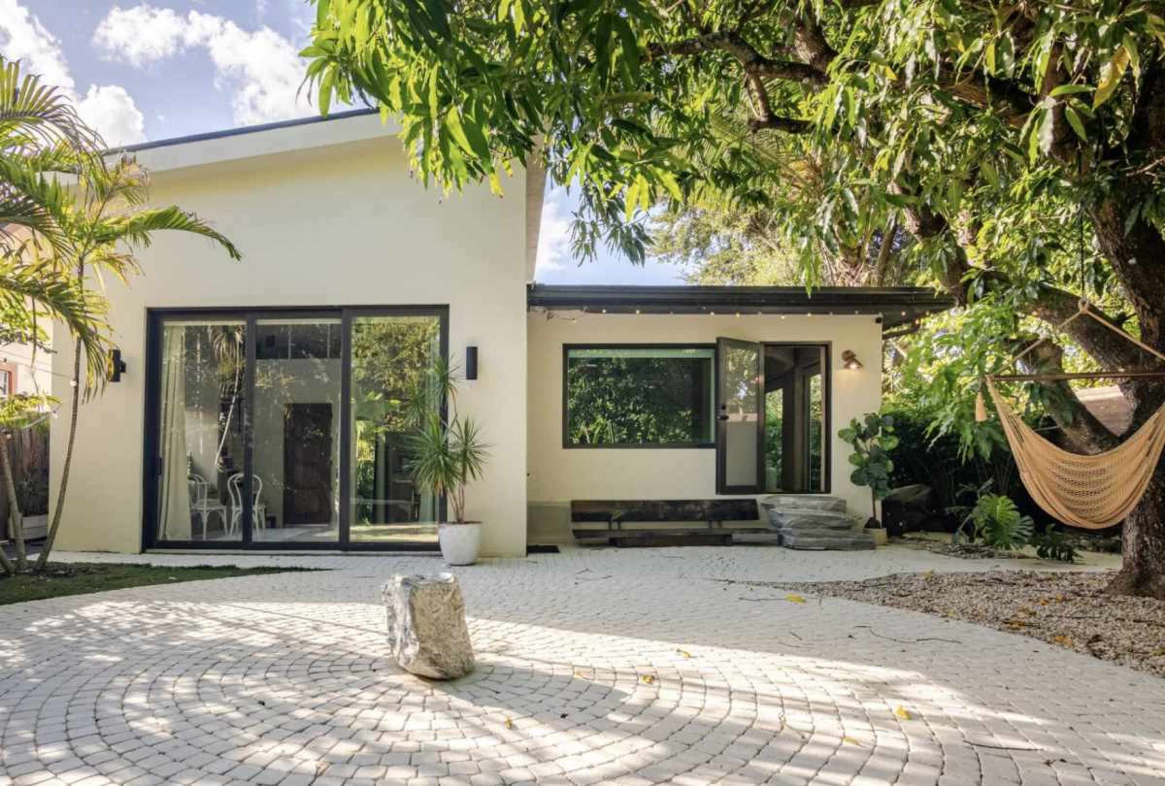 A modern home with large glass doors opens up to a landscaped garden featuring a circular stone pathway and a hammock beneath a tree.