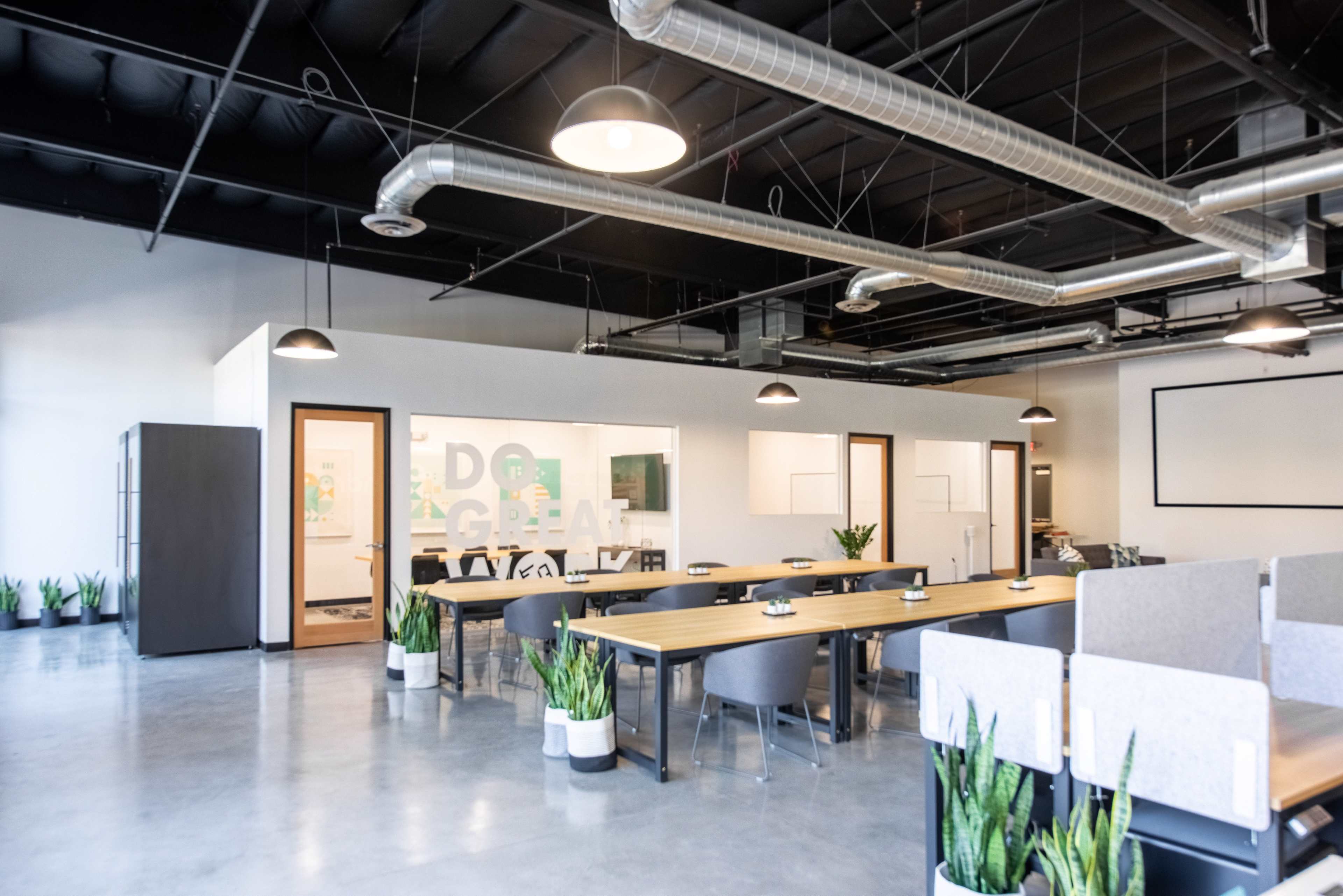 A modern office space featuring a large communal table, workstation dividers, and meeting rooms with glass walls, surrounded by potted plants.