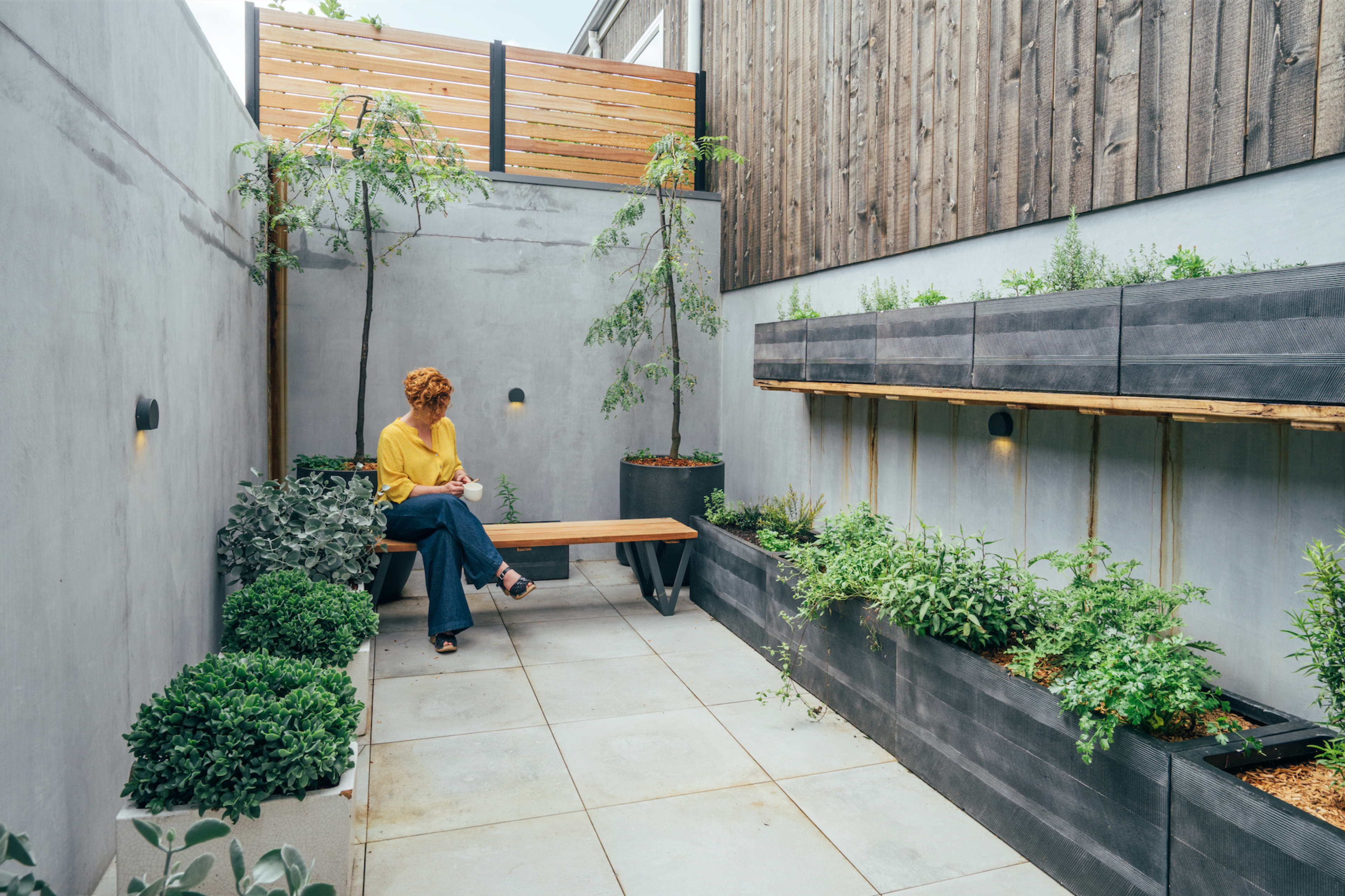 A person sits on a wooden bench in a narrow, modern garden space surrounded by concrete walls and raised planters filled with greenery.