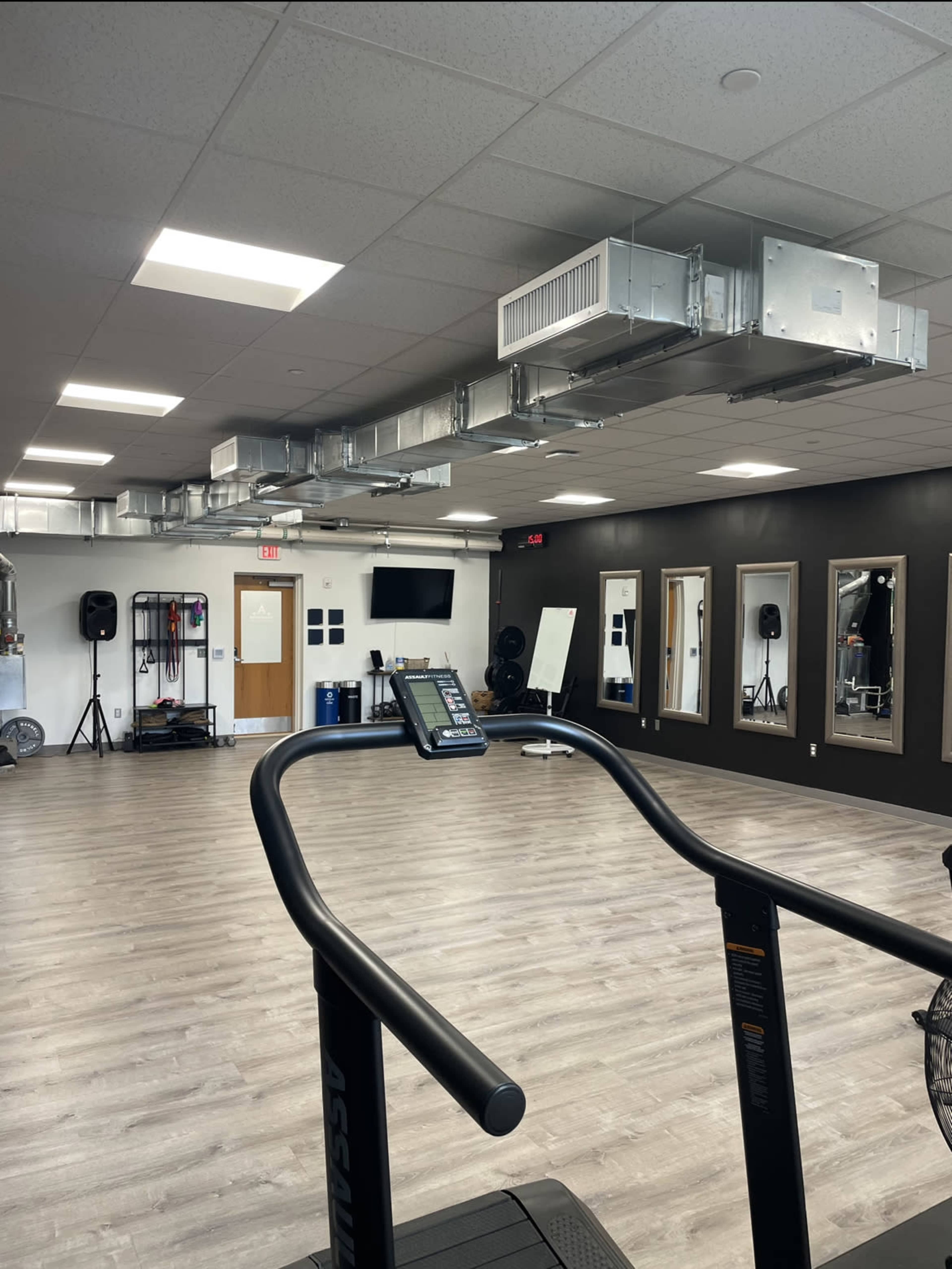A modern gym interior with a treadmill in the foreground, large mirrors on the walls, and ventilation ducts on the ceiling.