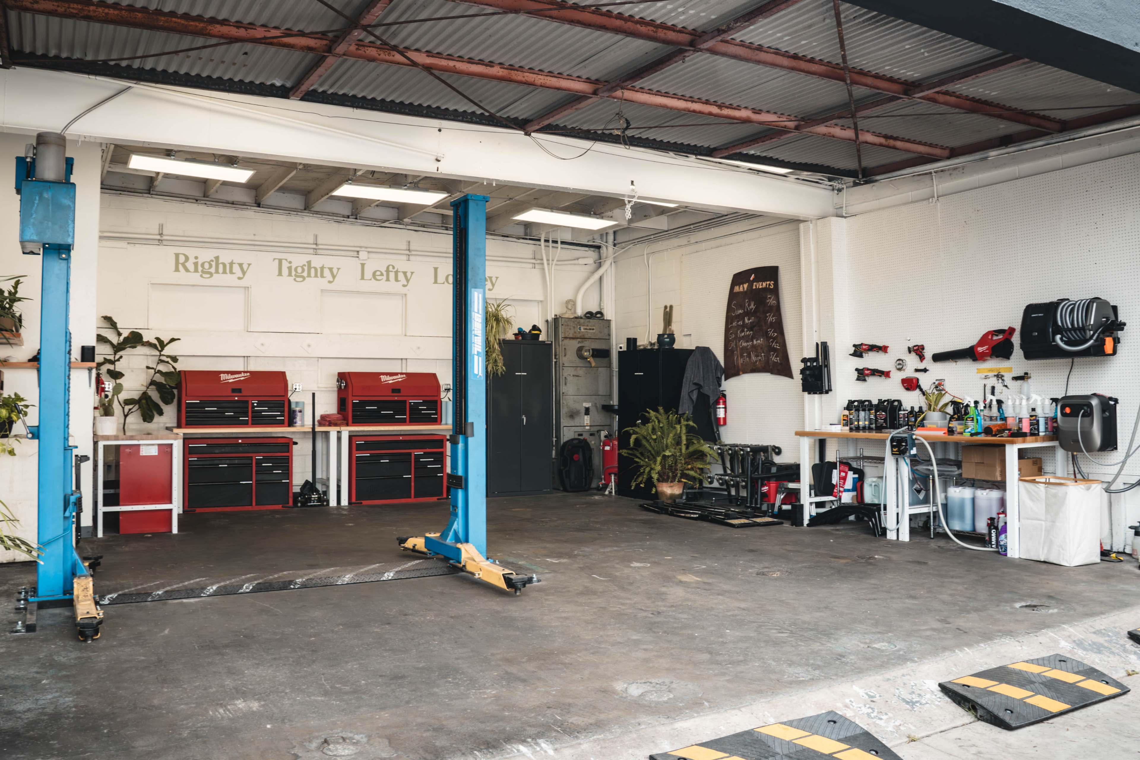 The image shows a spacious garage equipped with two blue car lifts, multiple workbenches, and tool storage units, along with potted plants and a wall-mounted tool organization system.