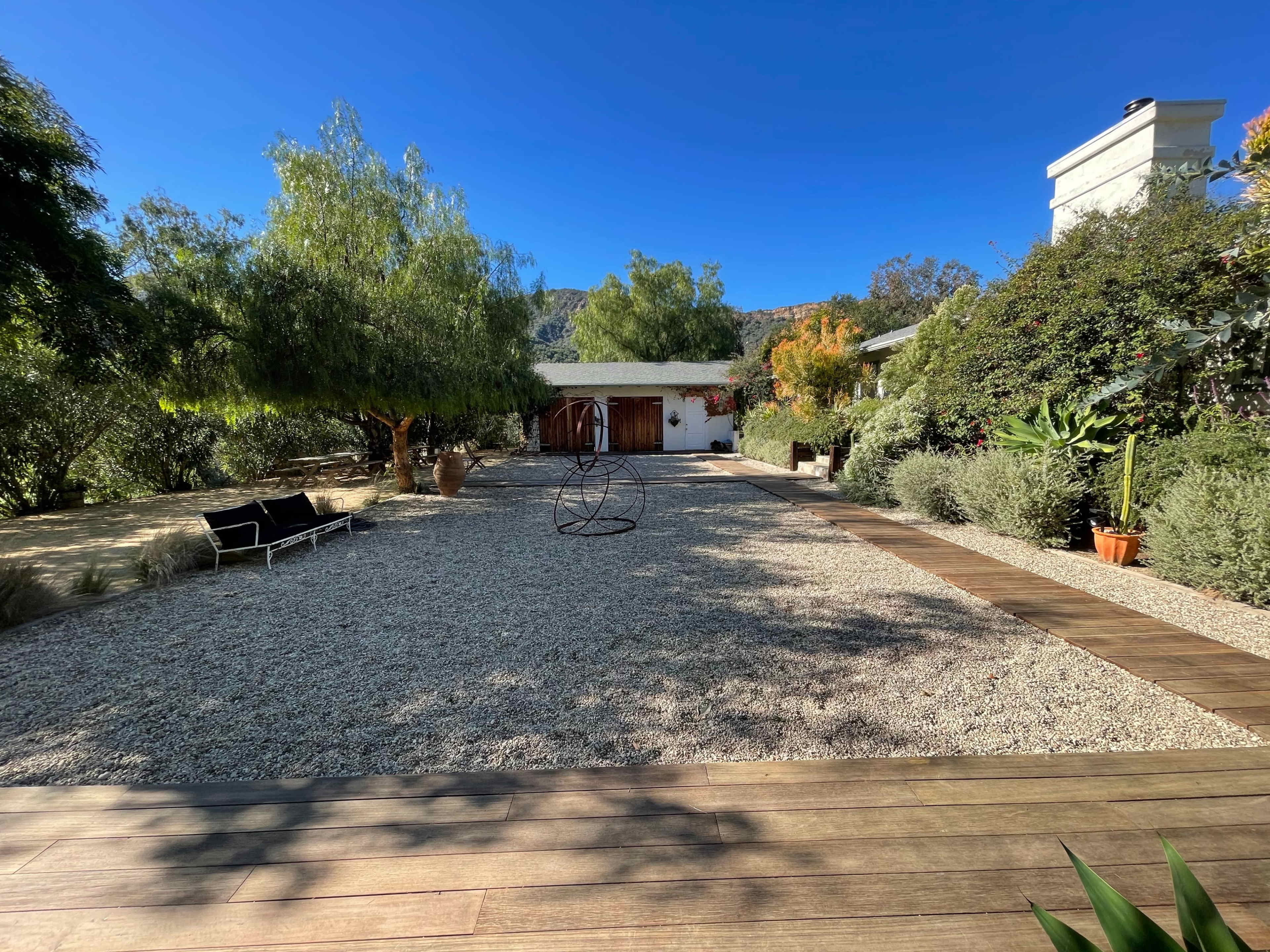 The image shows a landscaped outdoor area featuring gravel, wooden pathways, and a few trees, with a rustic building in the background.