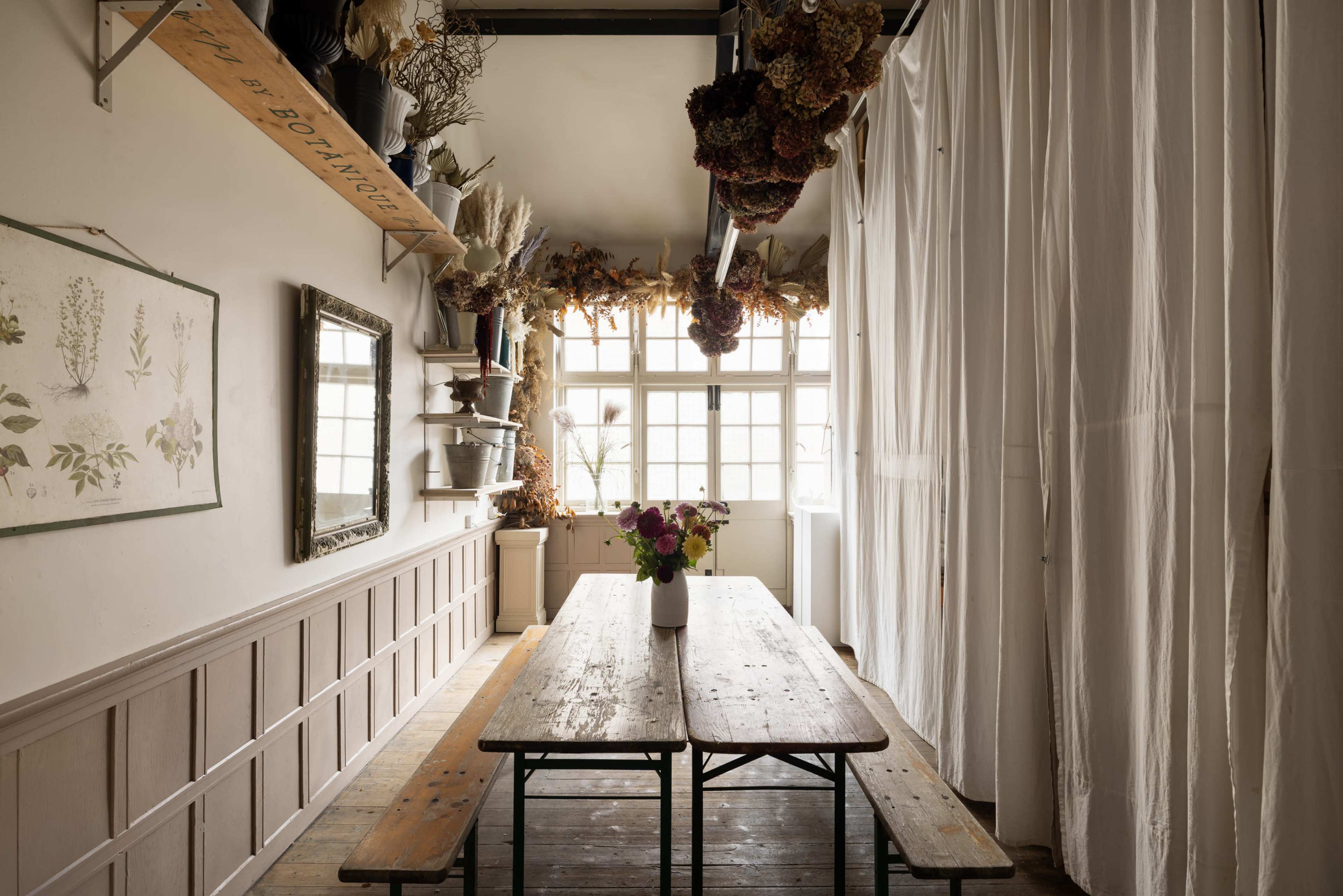 A rustic dining area features a wooden table with a vase of flowers, surrounded by floral decorations and soft, curtain-covered windows.