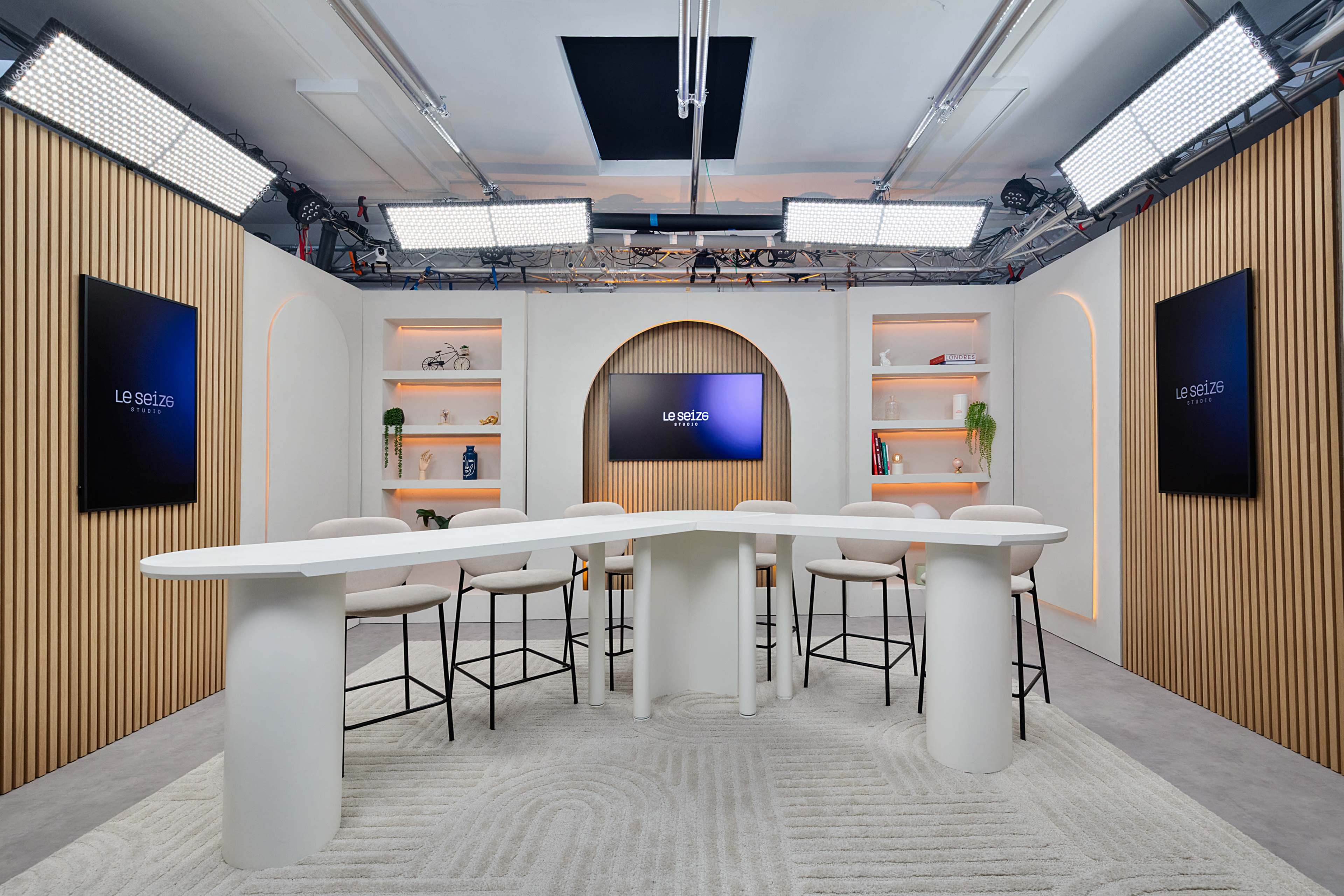 The image shows a modern studio set designed for a discussion or presentation, featuring a curved white table surrounded by several bar stools, with backlit shelves and screens on the walls.