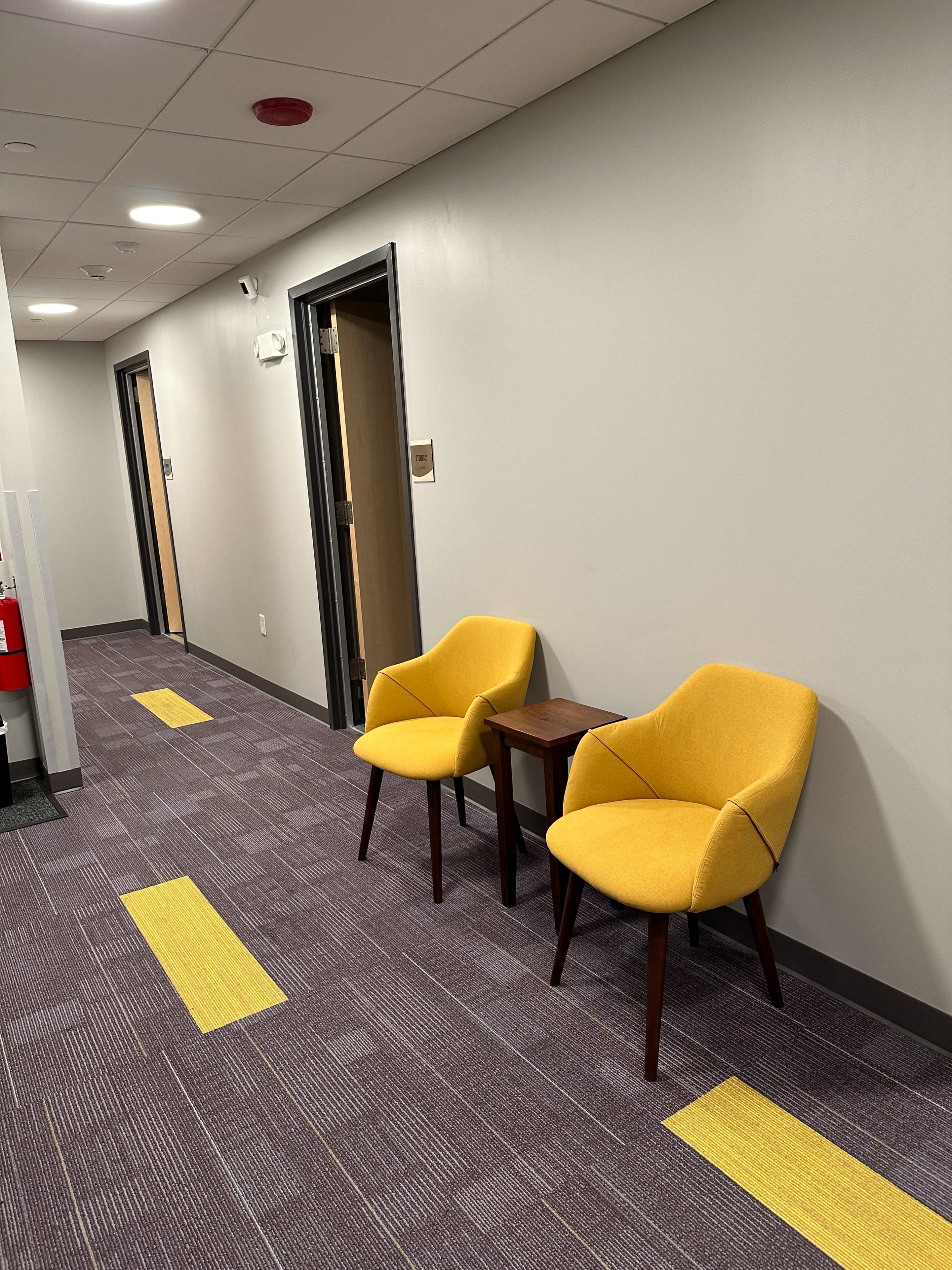 A hallway with two yellow chairs and a small table positioned against a wall, with doors along the corridor.