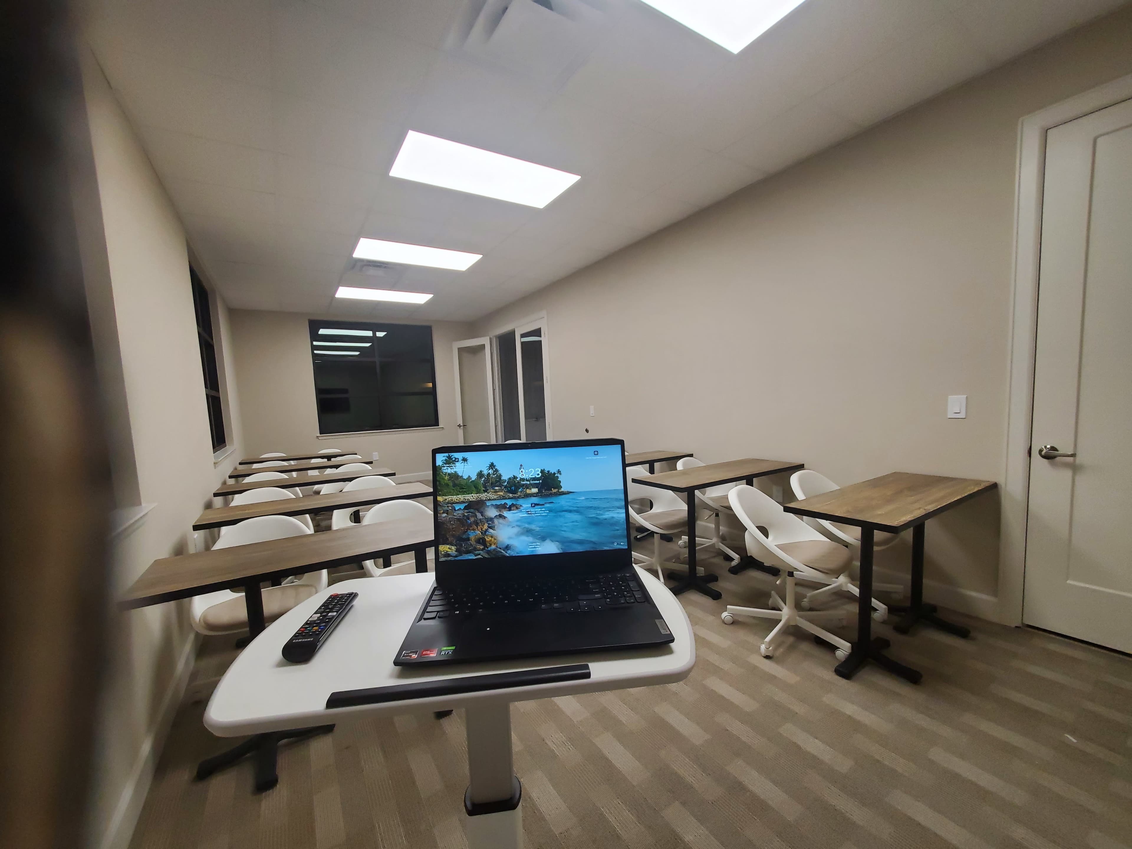 A laptop displaying a beach scene is set on a desk in a nearly empty classroom with rows of chairs and tables.