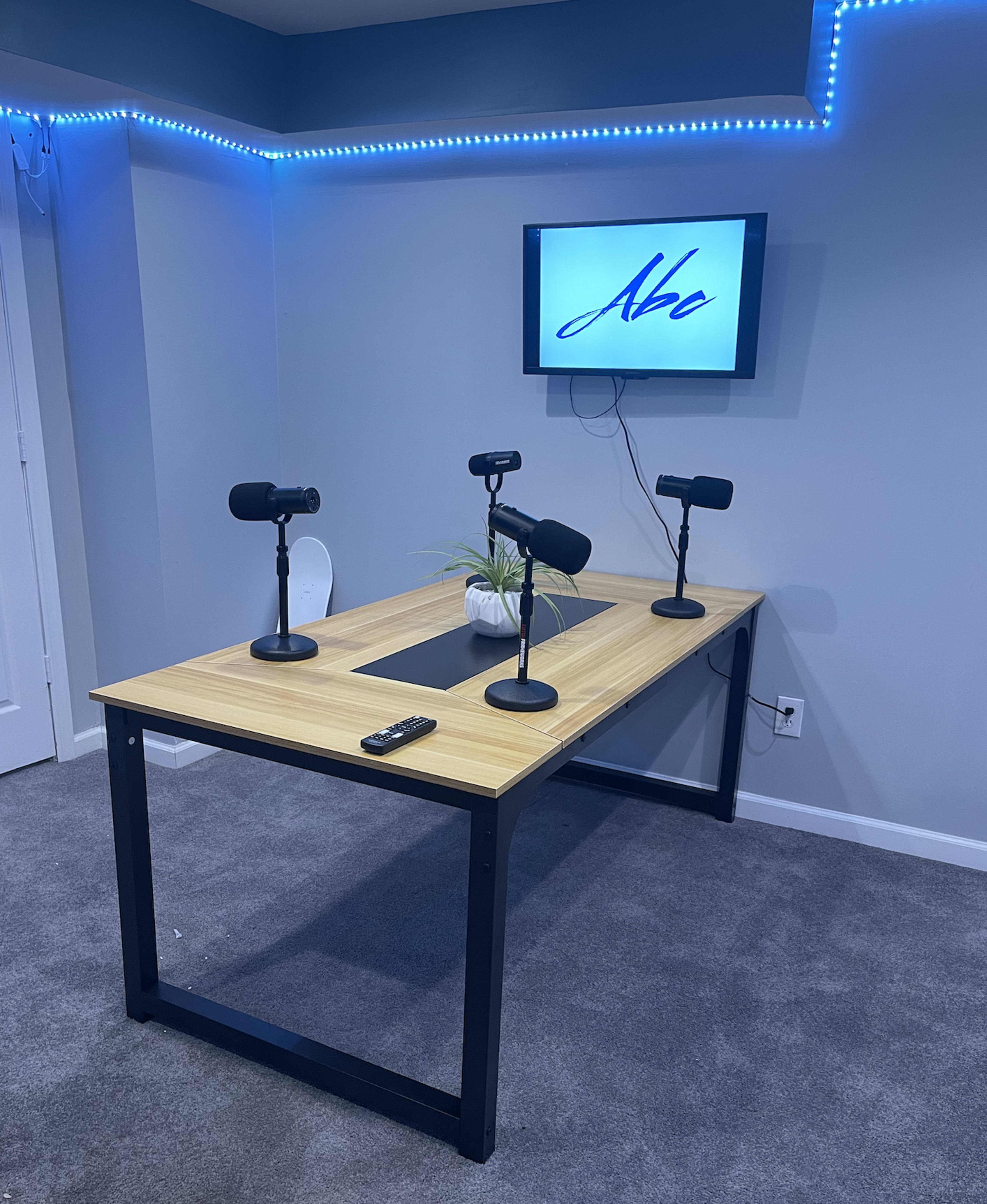 A podcasting setup with a wooden table, three microphones on stands, a small plant, and a screen displaying the text "Aba" mounted on a wall.