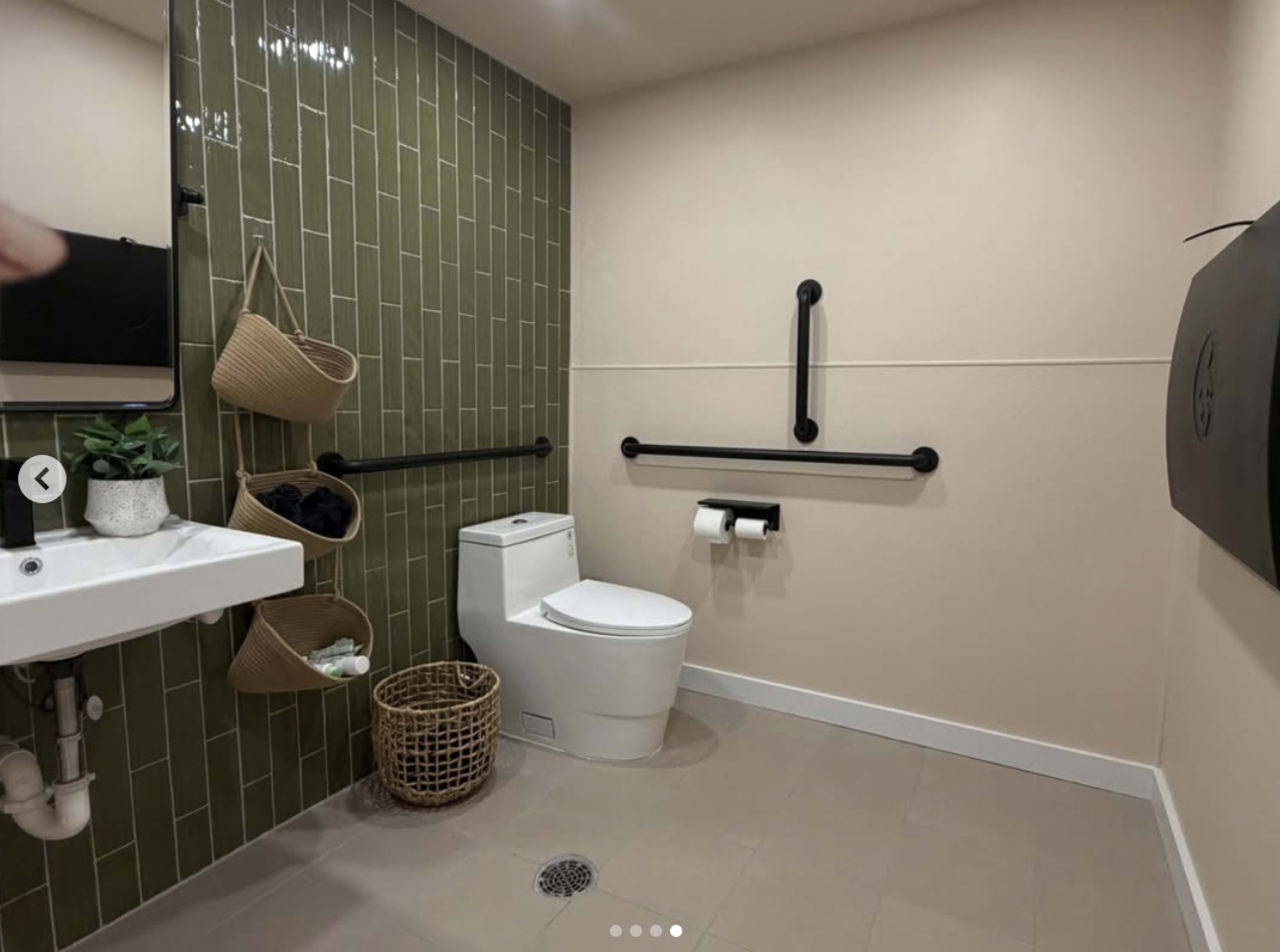 The image shows a restroom with a green tiled wall, a toilet, a sink with a basin, and accessories including handrails and baskets.
