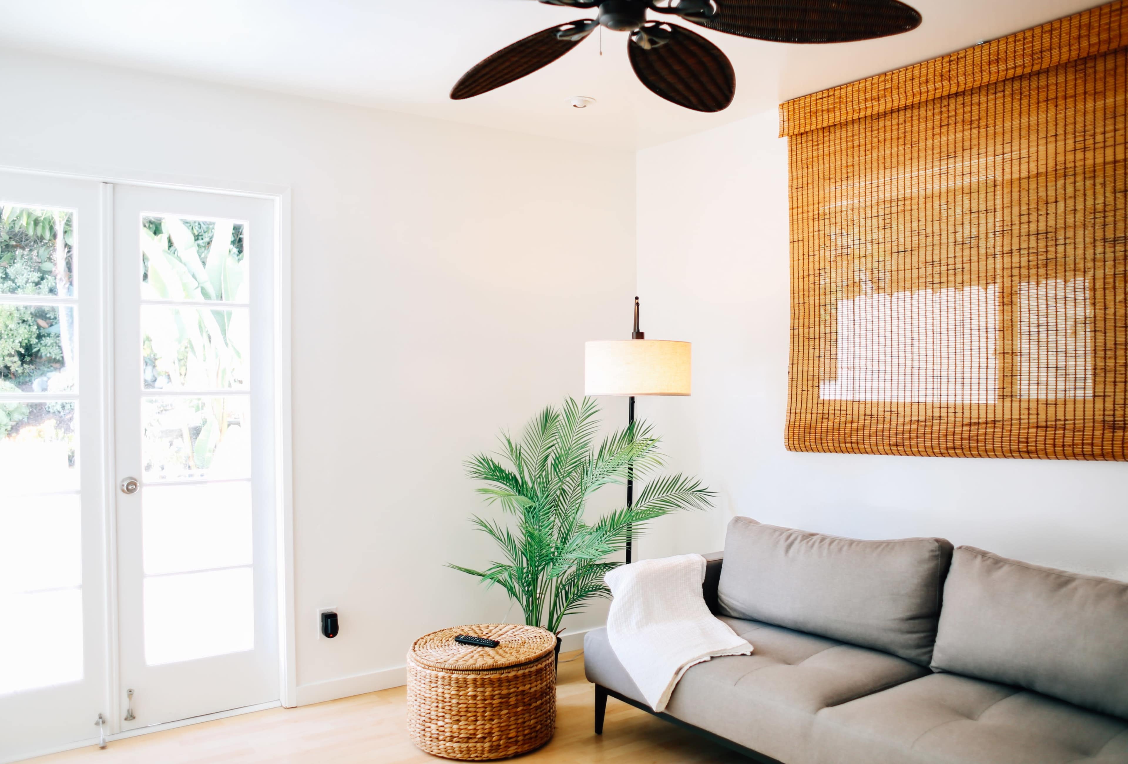 A light-filled living room features a gray sofa, a wicker basket table, a potted plant, and a ceiling fan above.