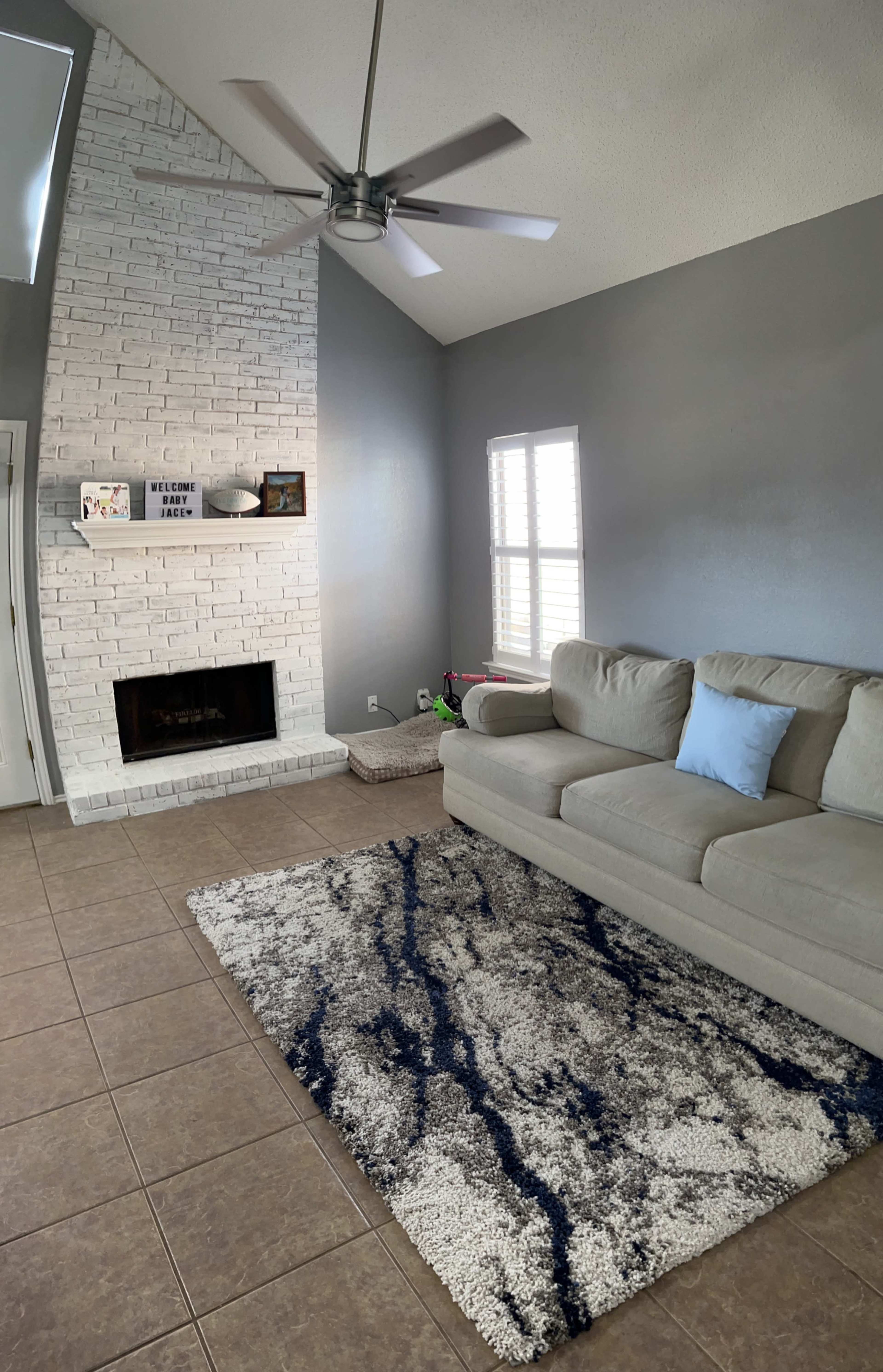 The image shows a living room with a sectional sofa, a textured rug, a brick fireplace, and a tall ceiling with a ceiling fan.