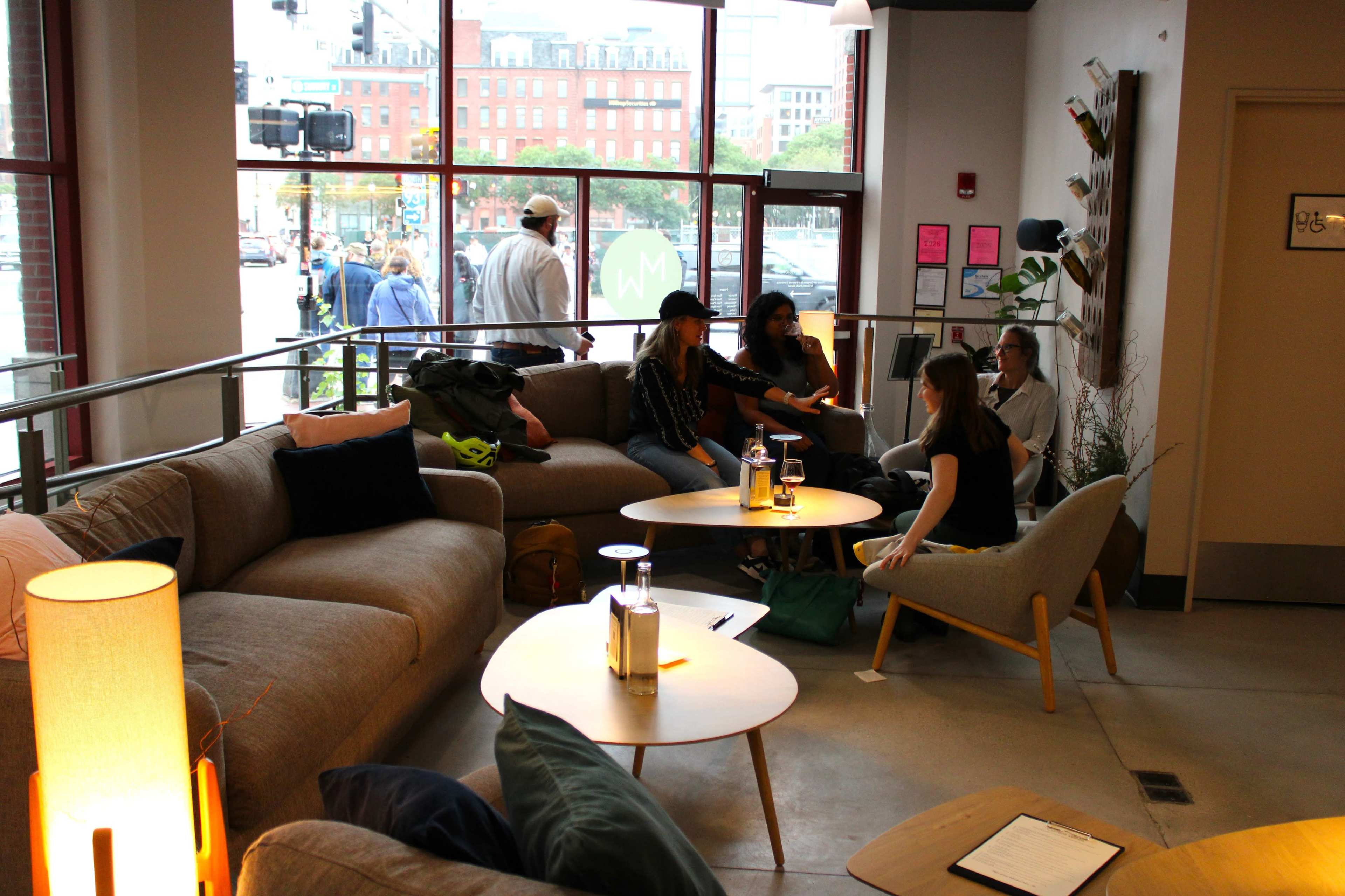 A group of four people sit around a coffee table in a modern lounge area, with large windows overlooking a city street.