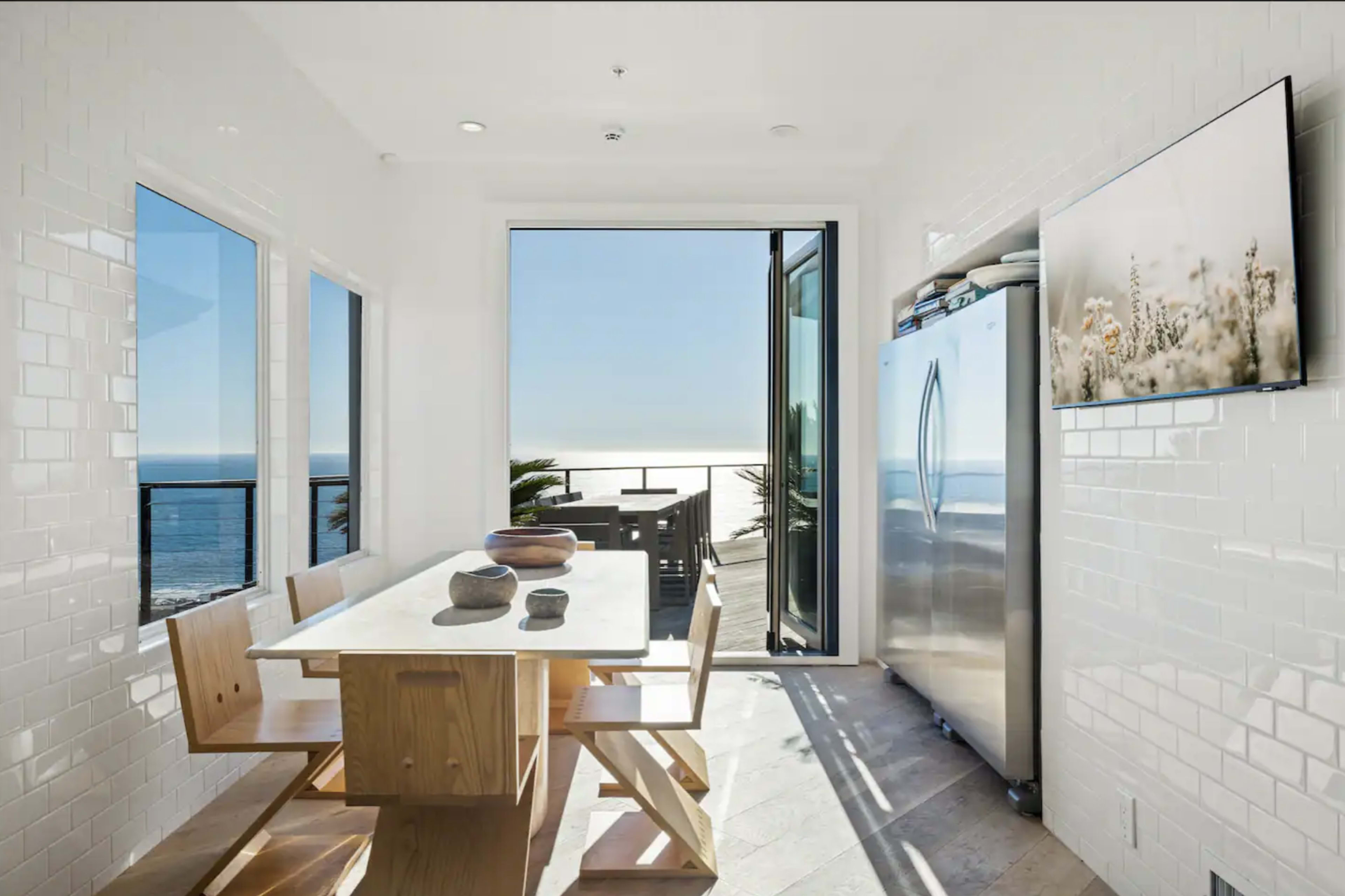 A bright dining area features a wooden table and chairs, with large windows overlooking the ocean.