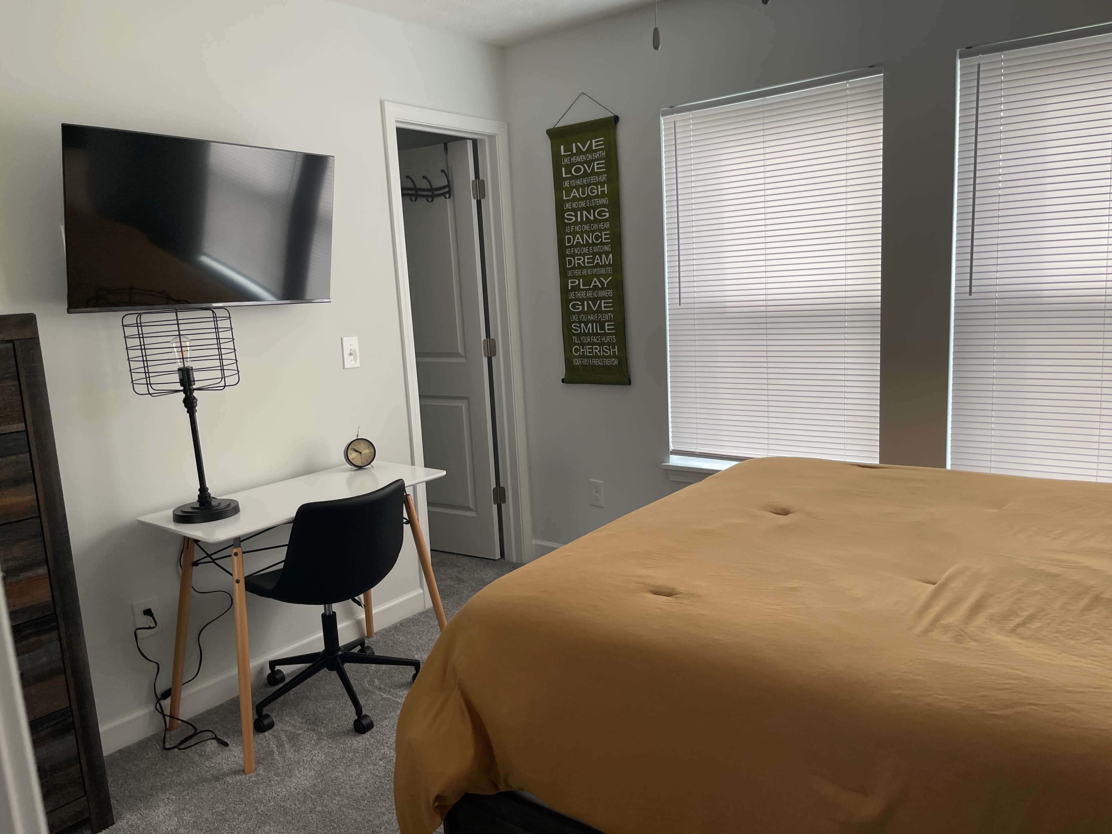 A bedroom with a bed covered in a yellow blanket, a desk with a chair and clock, a wall-mounted television, and two windows with blinds.