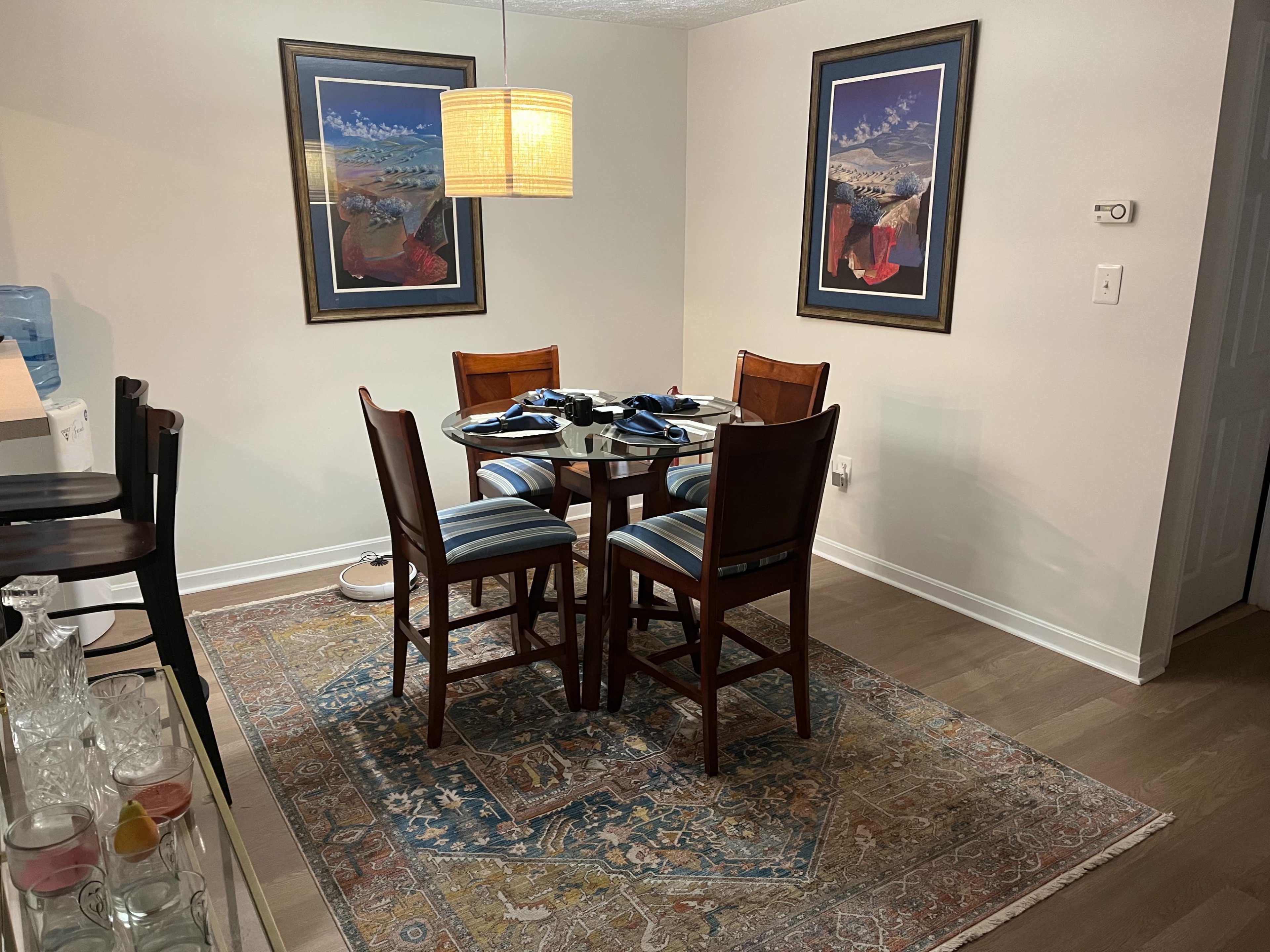 A dining area with a glass-top table surrounded by four wooden chairs, a patterned rug on the floor, and framed artwork on the walls.