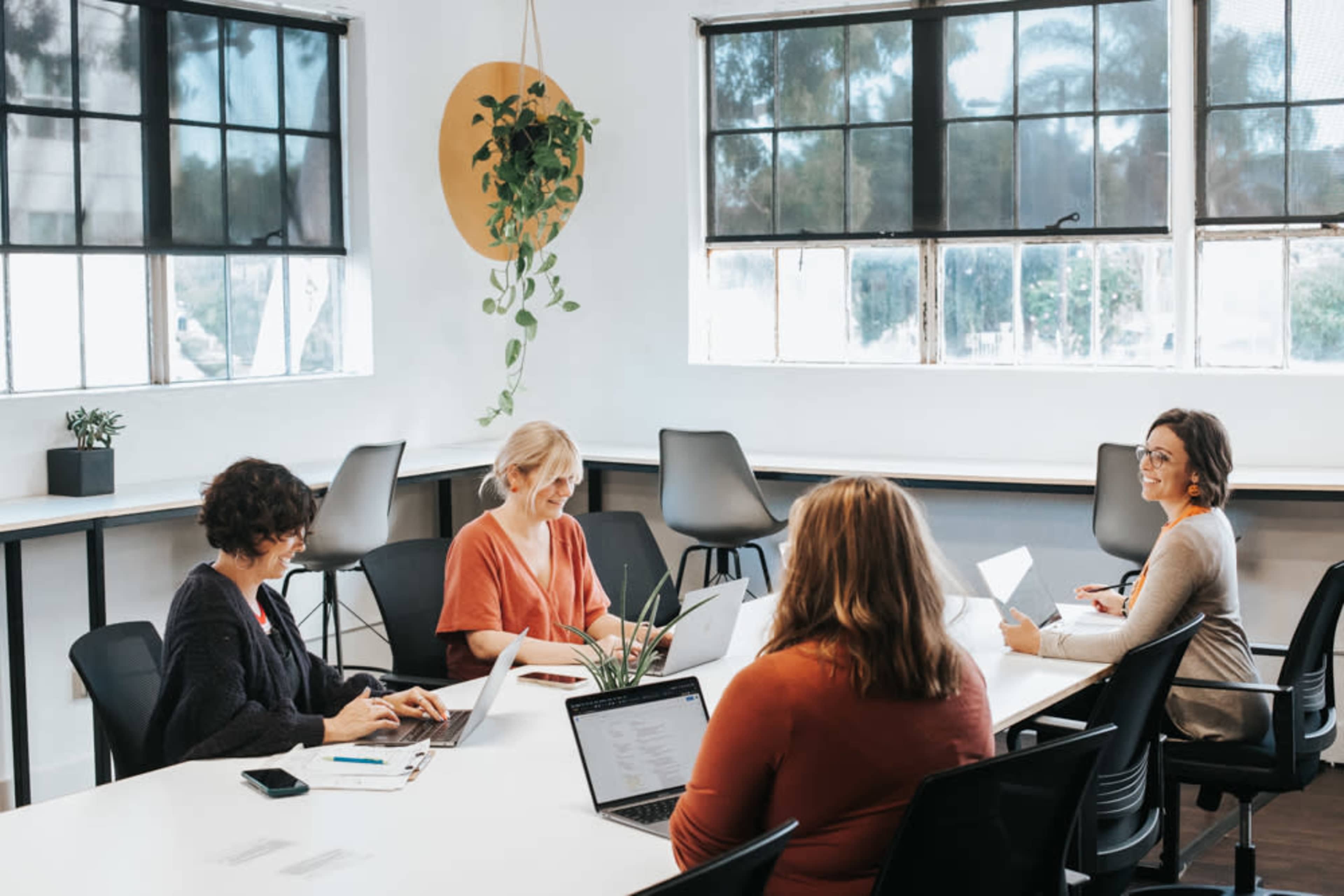 A group of five people are seated around a table in a well-lit office space, working on laptops and notebooks.
