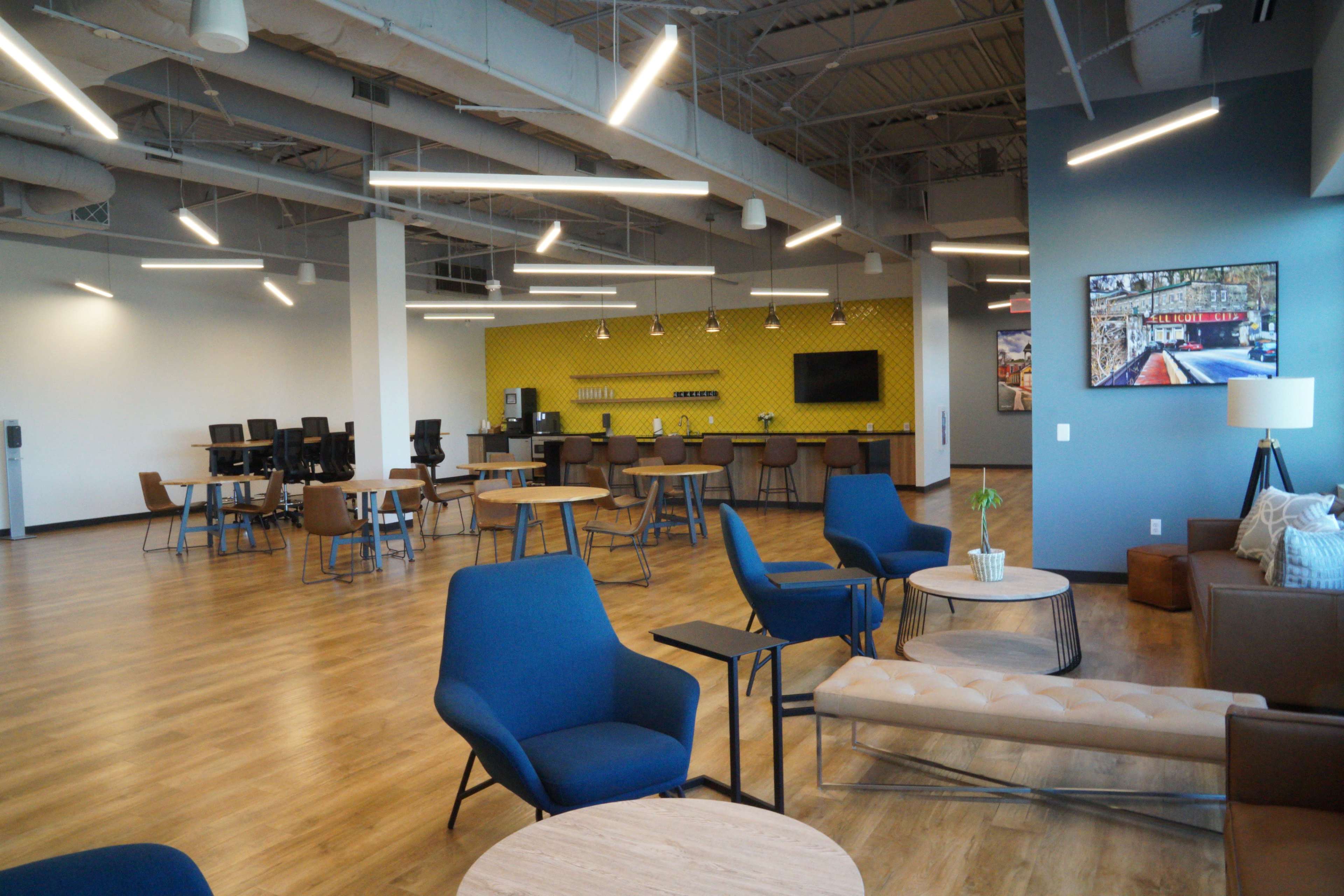 The image shows a modern, open workspace featuring wooden floors, blue and yellow accent walls, and various seating arrangements with tables.