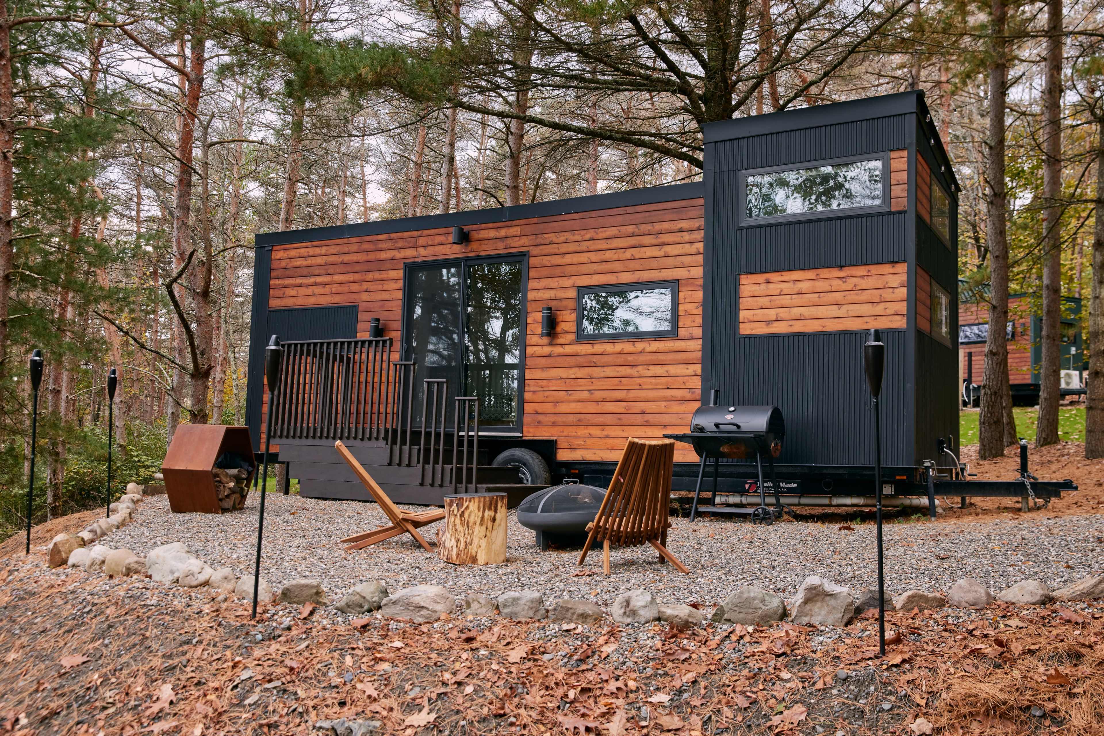 A modern tiny house with a wooden exterior is situated in a forested area, surrounded by gravel and outdoor seating.