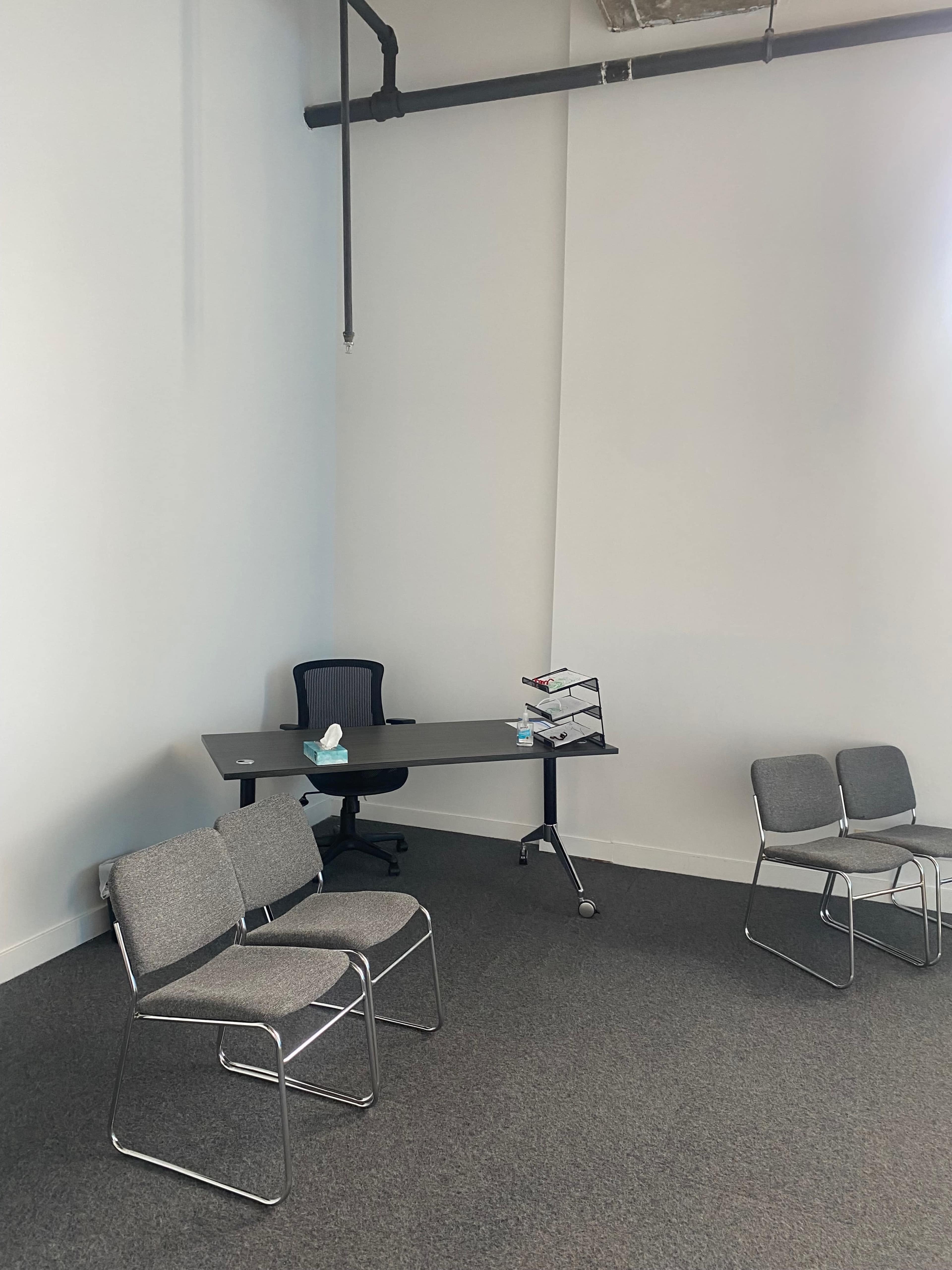 A sparsely furnished office space with a desk, a chair, and four empty gray chairs arranged around a small area.