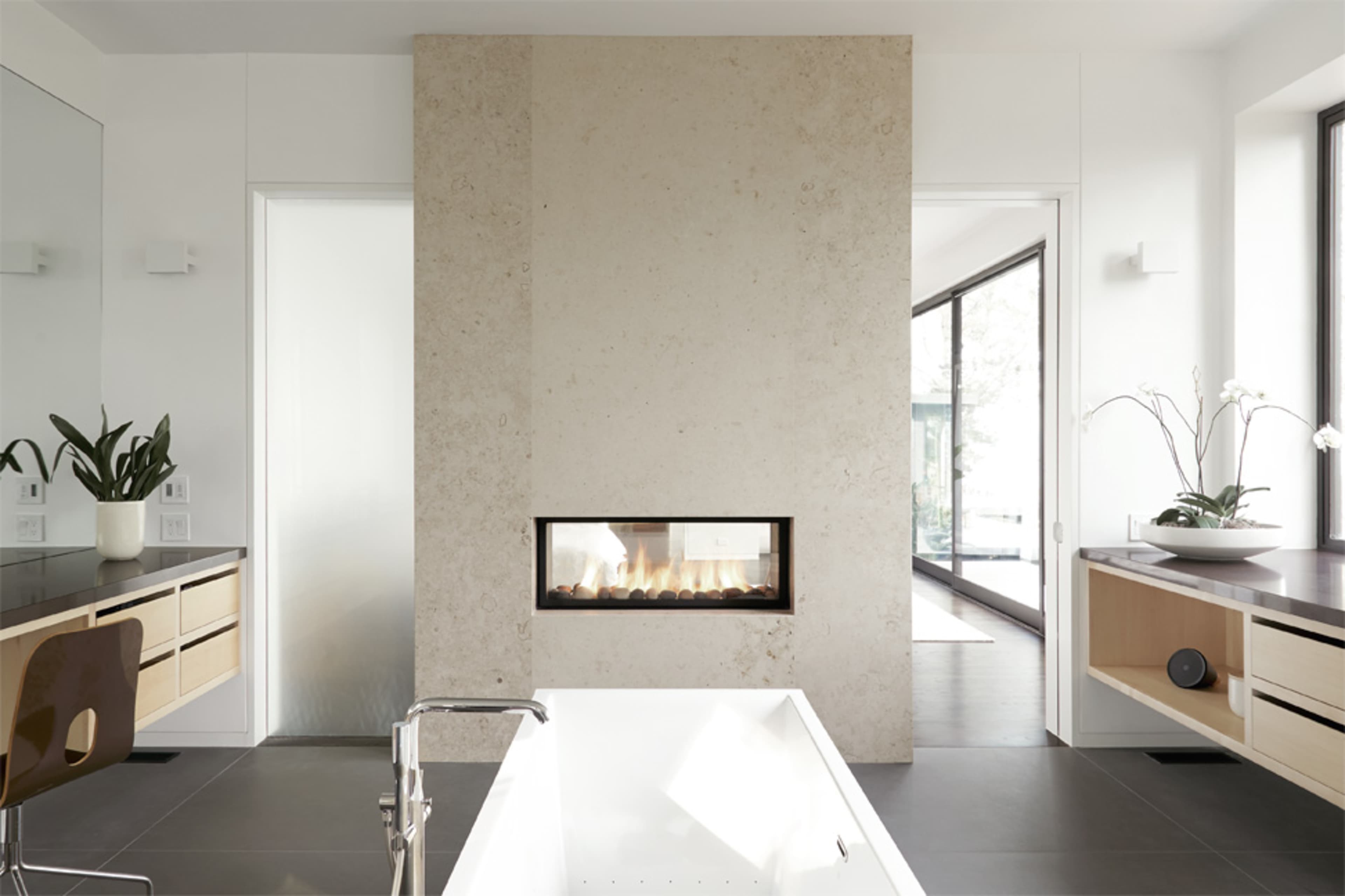 A modern bathroom features a freestanding bathtub, a minimalist fireplace embedded in a textured wall, and large windows allowing natural light.