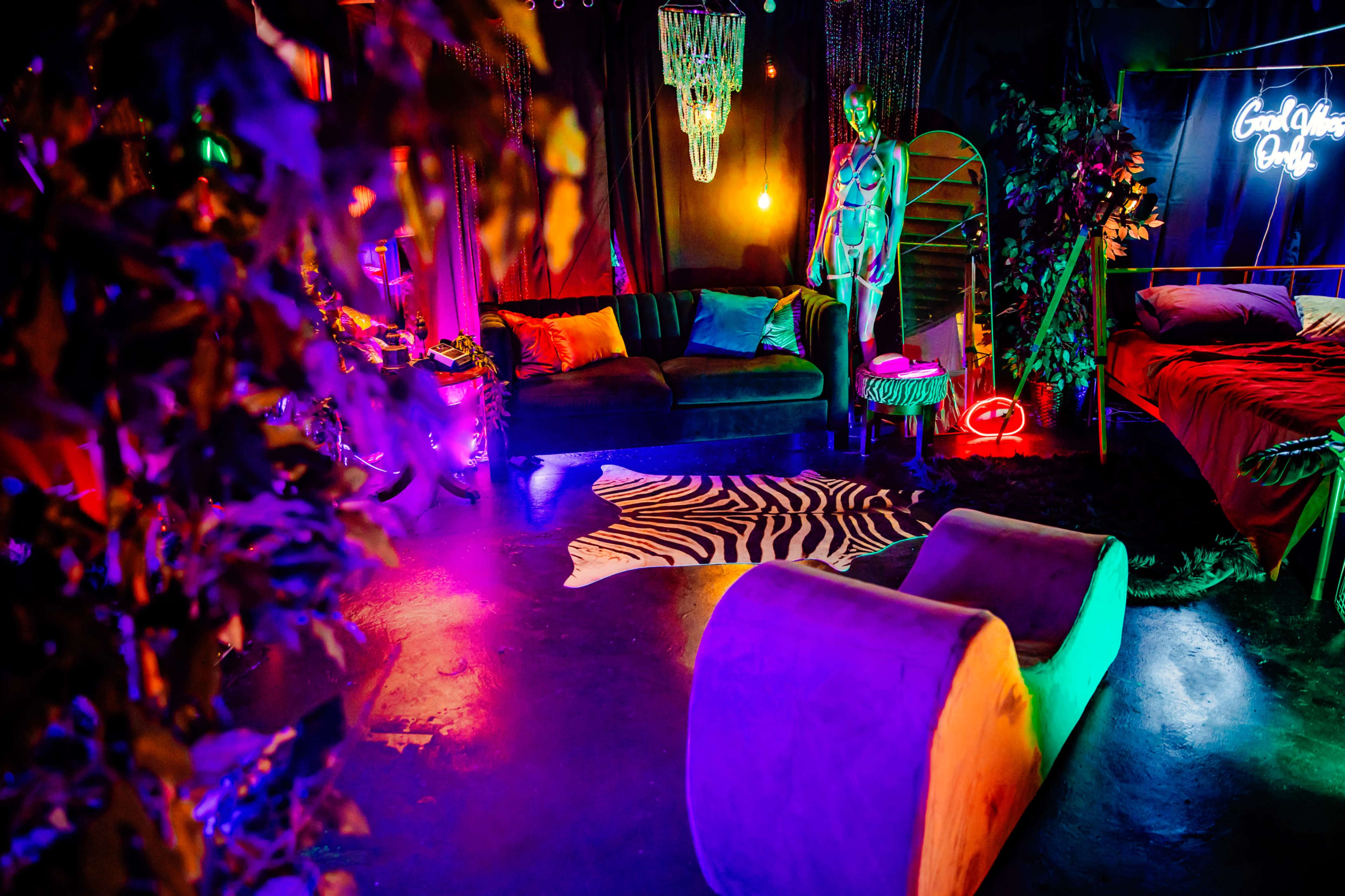The image depicts a vibrant, dimly lit room with colorful lighting, featuring a couch, a bed, a zebra-striped rug, and a mannequin.