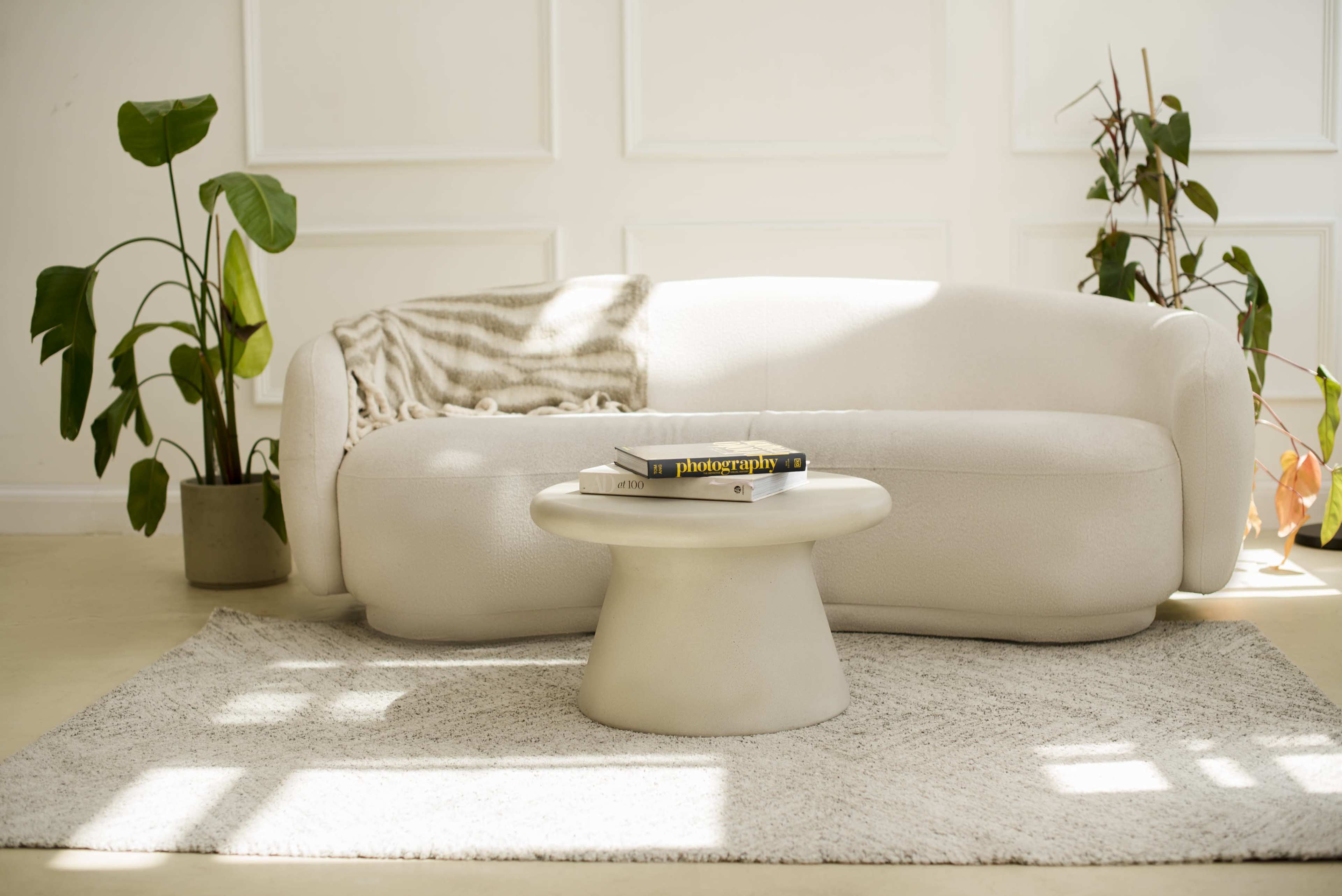 A light-colored sofa is positioned next to a round, minimalist coffee table on a textured rug, with sunlight casting shadows across the scene.