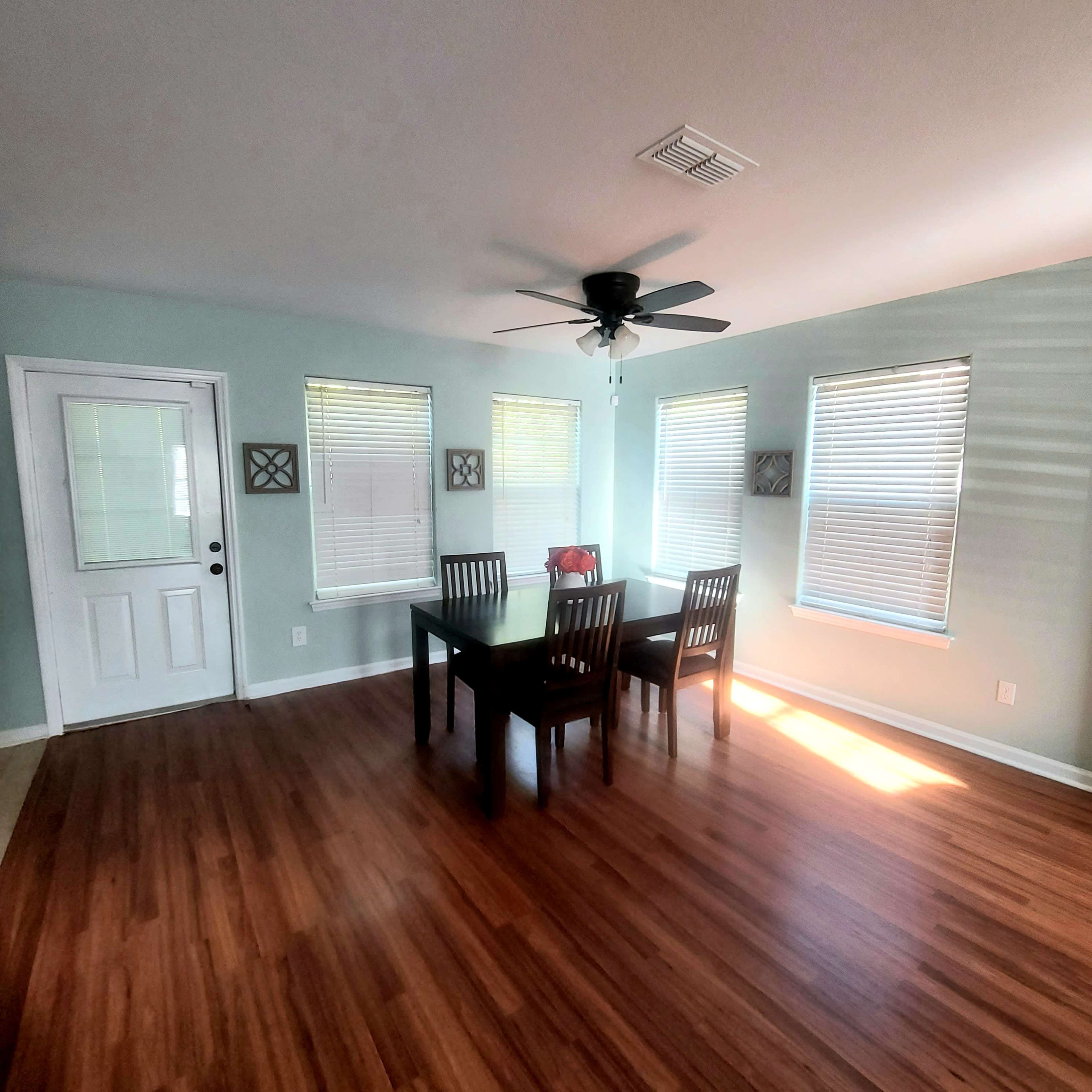 A dining area features a table with four chairs, a ceiling fan, and several windows with blinds, all set against light green walls and hardwood flooring.